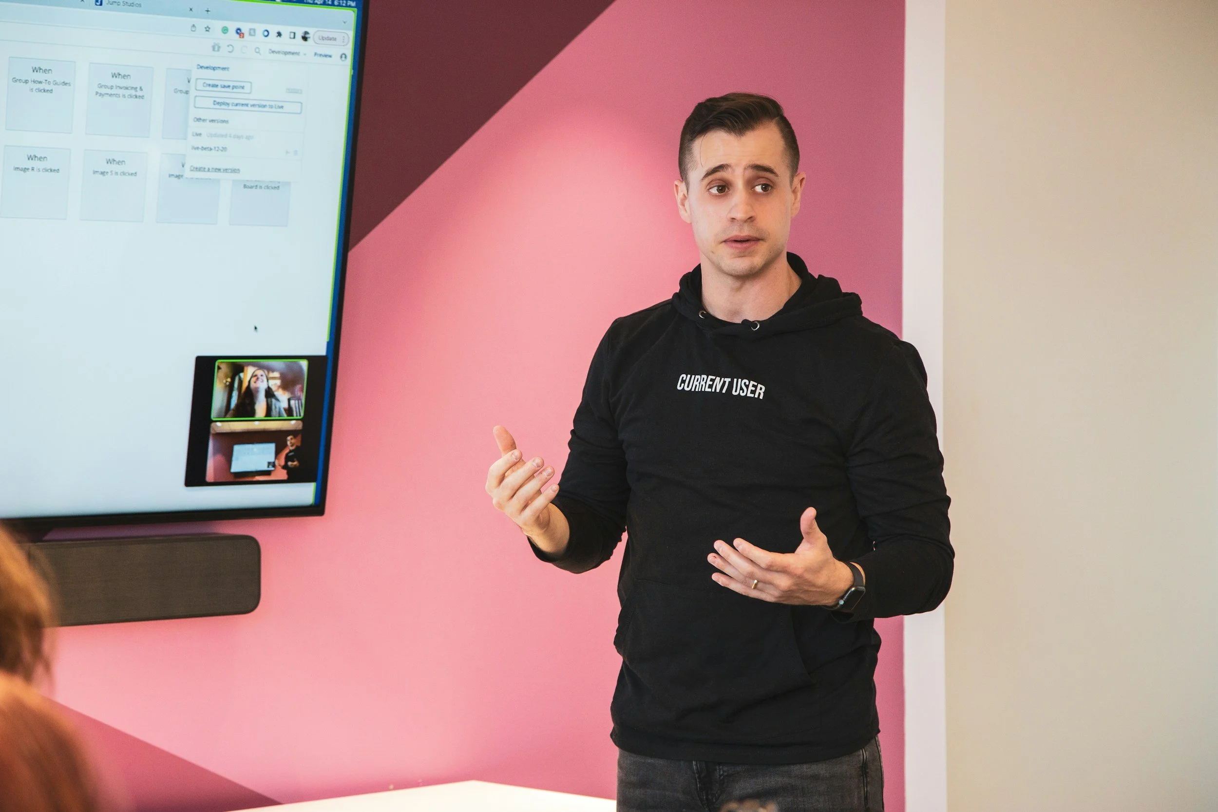A man in a black shirt holding a notebook and pointing while speaking to a group outdoors on a sunny day, with a soccer goal and houses in the background.