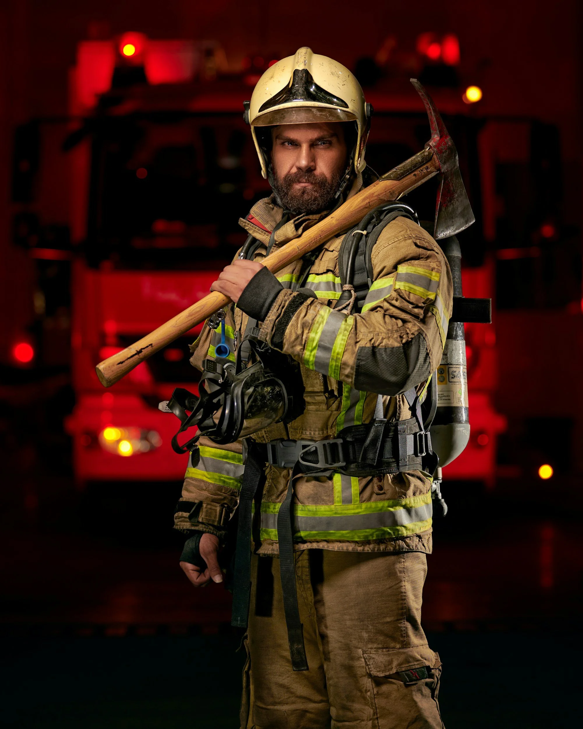 Firefighter standing on the street, holding a tool, with fire trucks in the background.