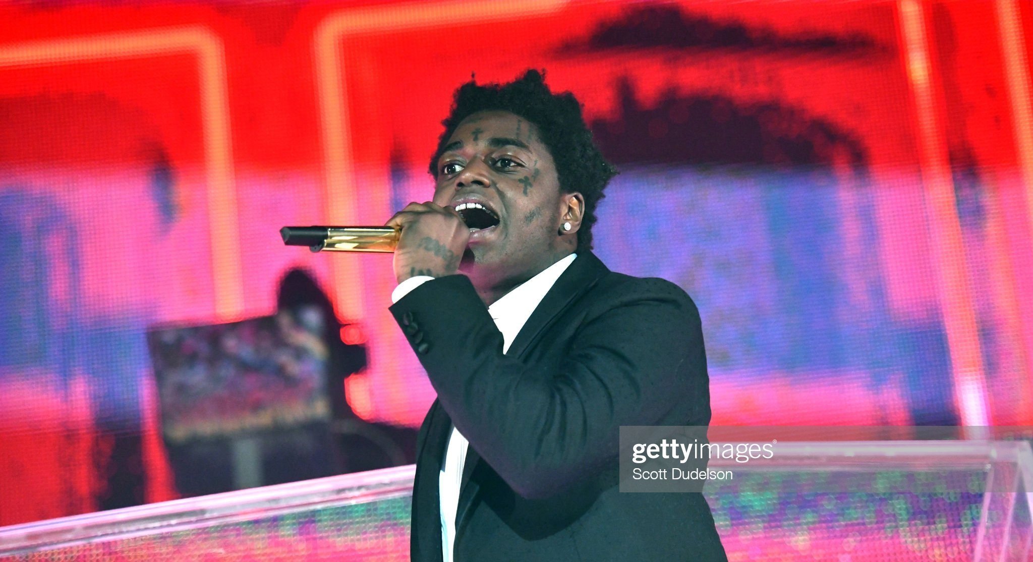A man singing into a gold microphone on stage, dressed in a black suit and white shirt, with a vibrant pink and purple blurred digital background.