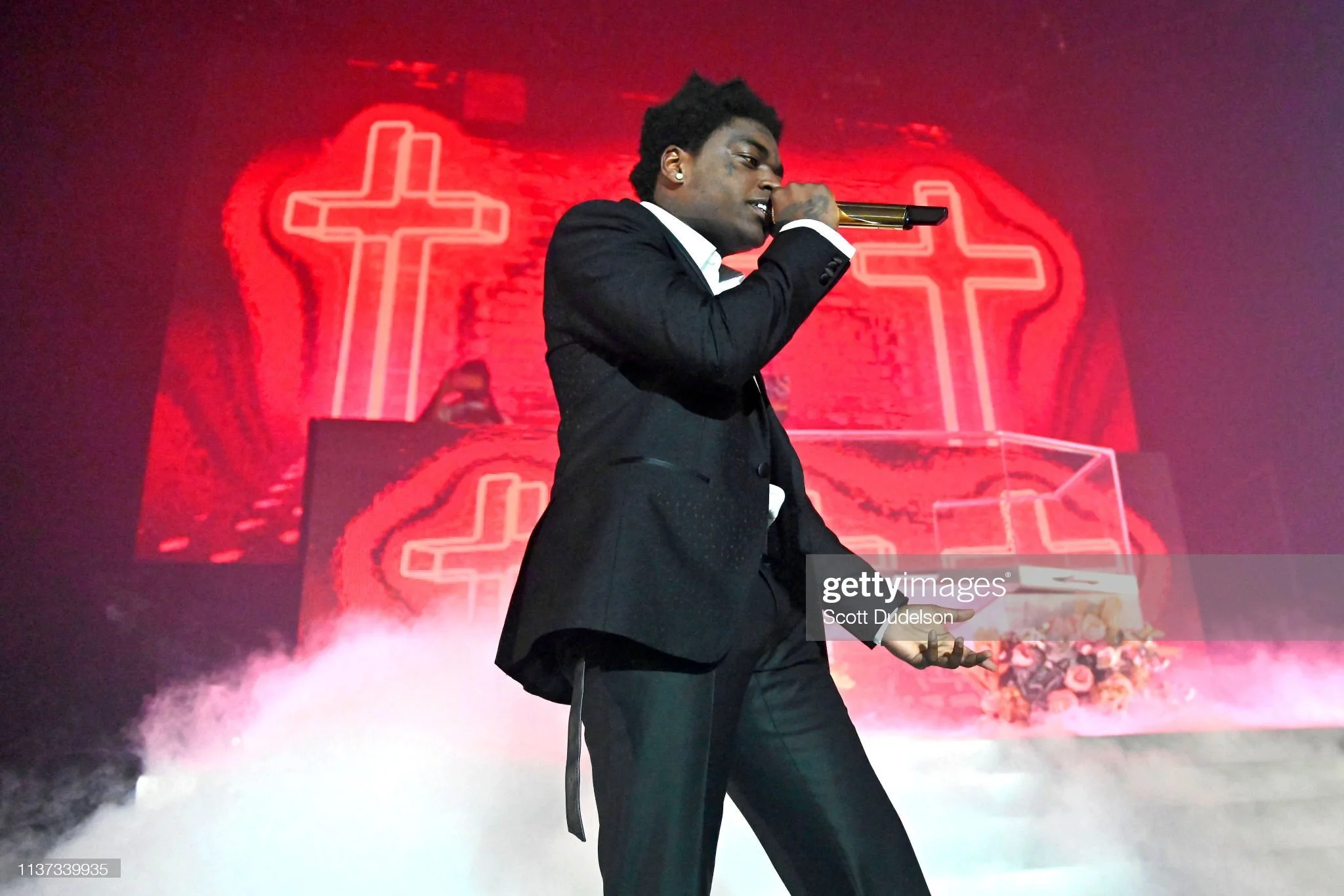 A man in a black suit performs on stage, holding a gold microphone. Behind him, there is a red neon cross and a similar outline, creating a striking visual backdrop. The stage is filled with mist, enhancing the dramatic effect.