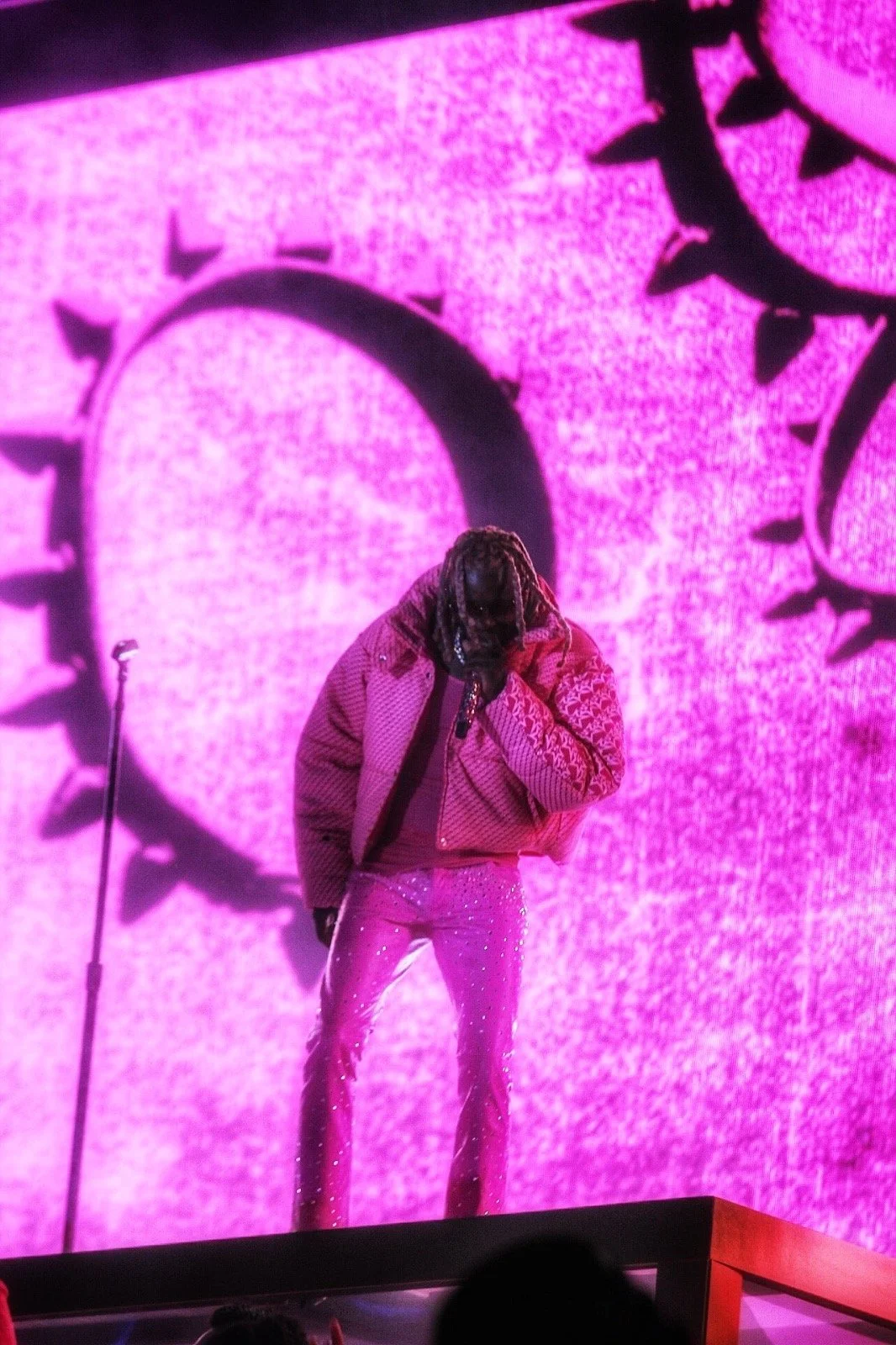 A performer stands on stage with a pink background featuring a large clock face with Roman numerals and spikes. The performer is dressed in a pink jacket and pants with sparkles, covering their face with their hand.