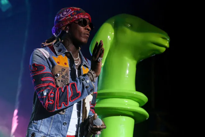 Person with dreadlocks wearing a colorful hat, sunglasses, and a denim jacket with patches, standing next to a large green sculpture of a hand making a peace sign.
