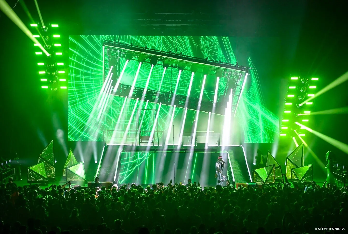 A concert stage with vibrant green laser lights and geometric LED structures, a large digital screen displaying green grid-like patterns, and an audience watching the performance.