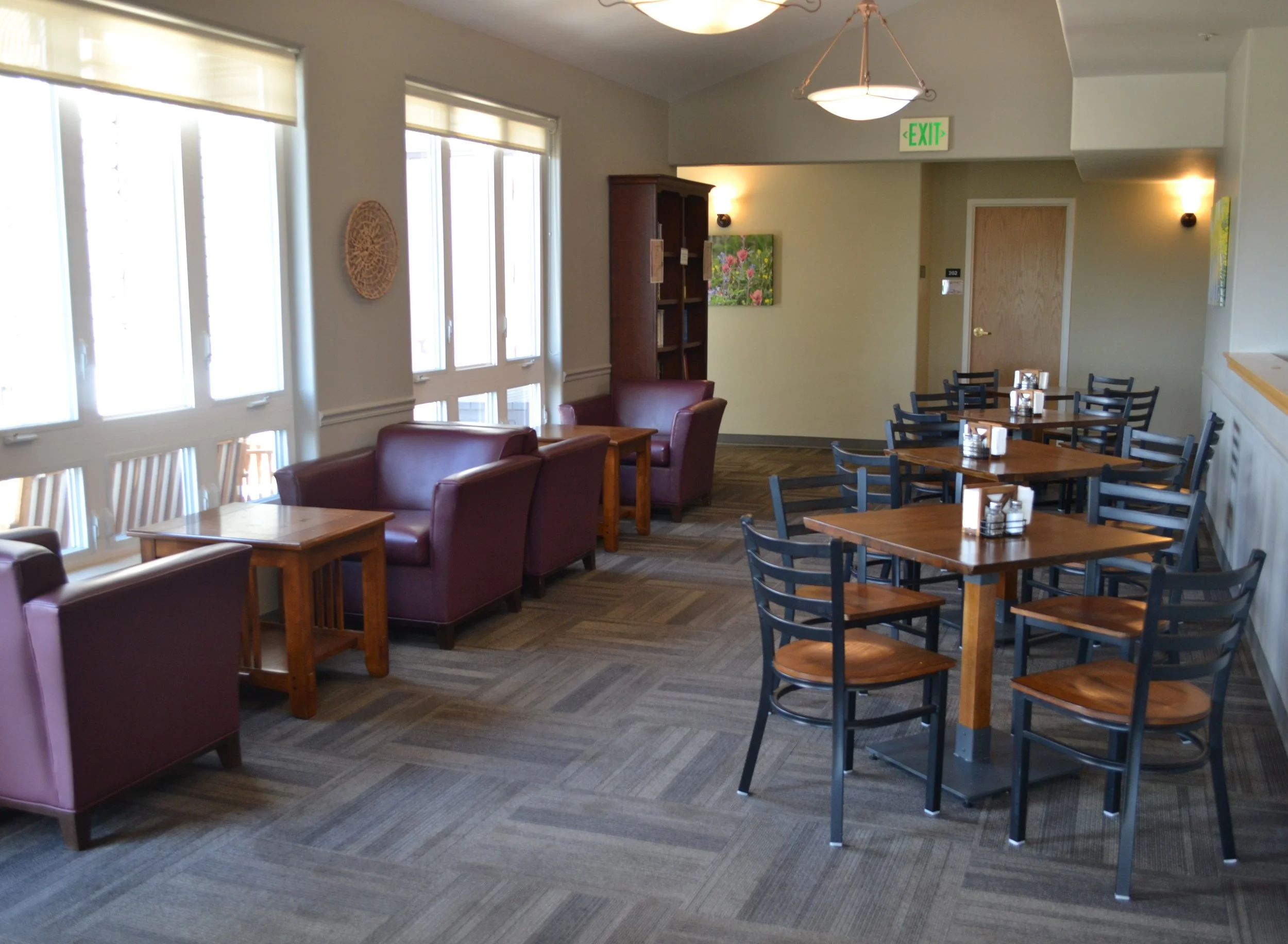 A cozy cafe interior with tables and chairs, maroon armchairs, and large windows.