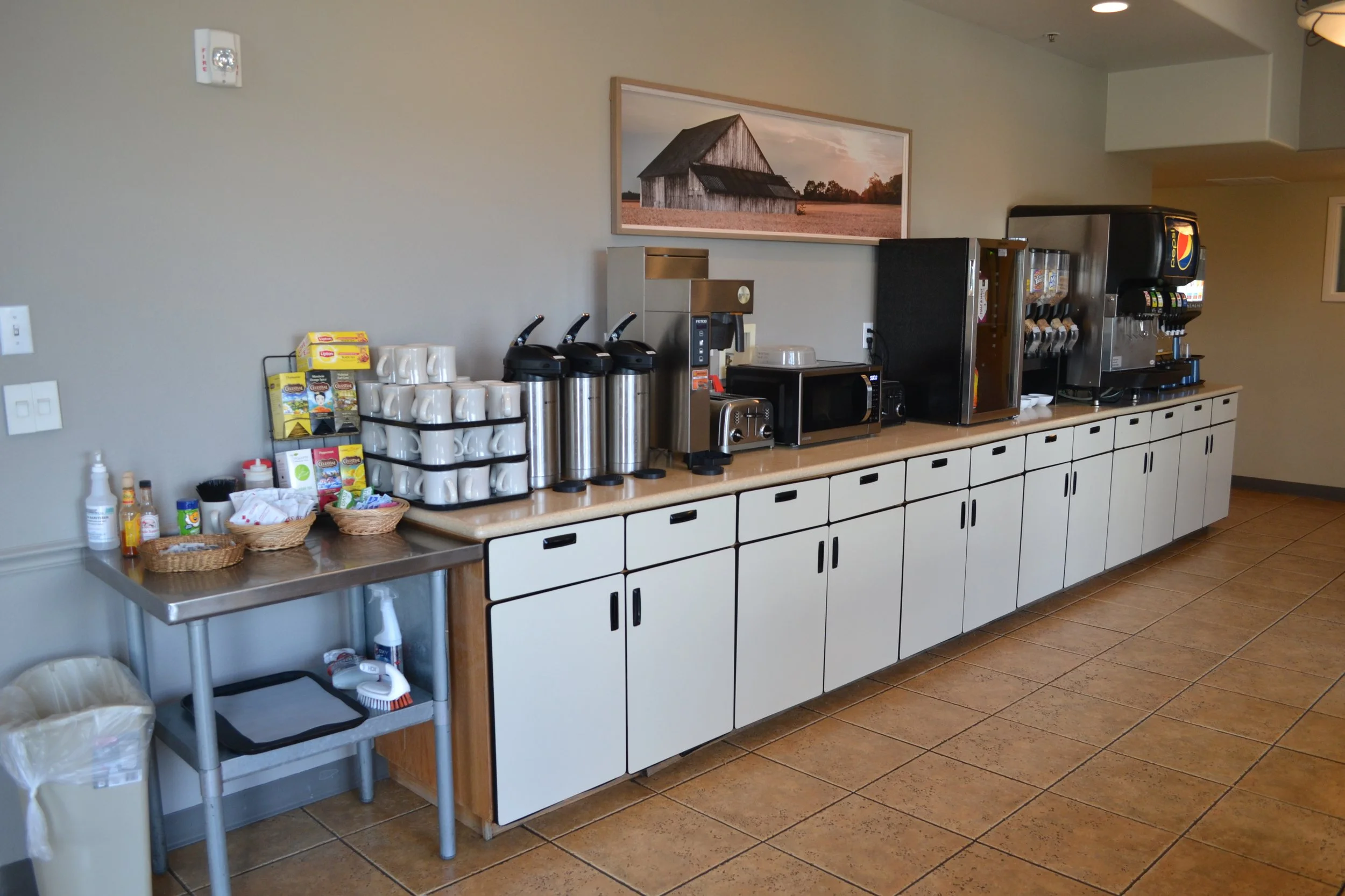 Breakfast area with coffee and tea station, beverage dispenser, microwave, toaster, and condiments on countertop in a room with tiled flooring.