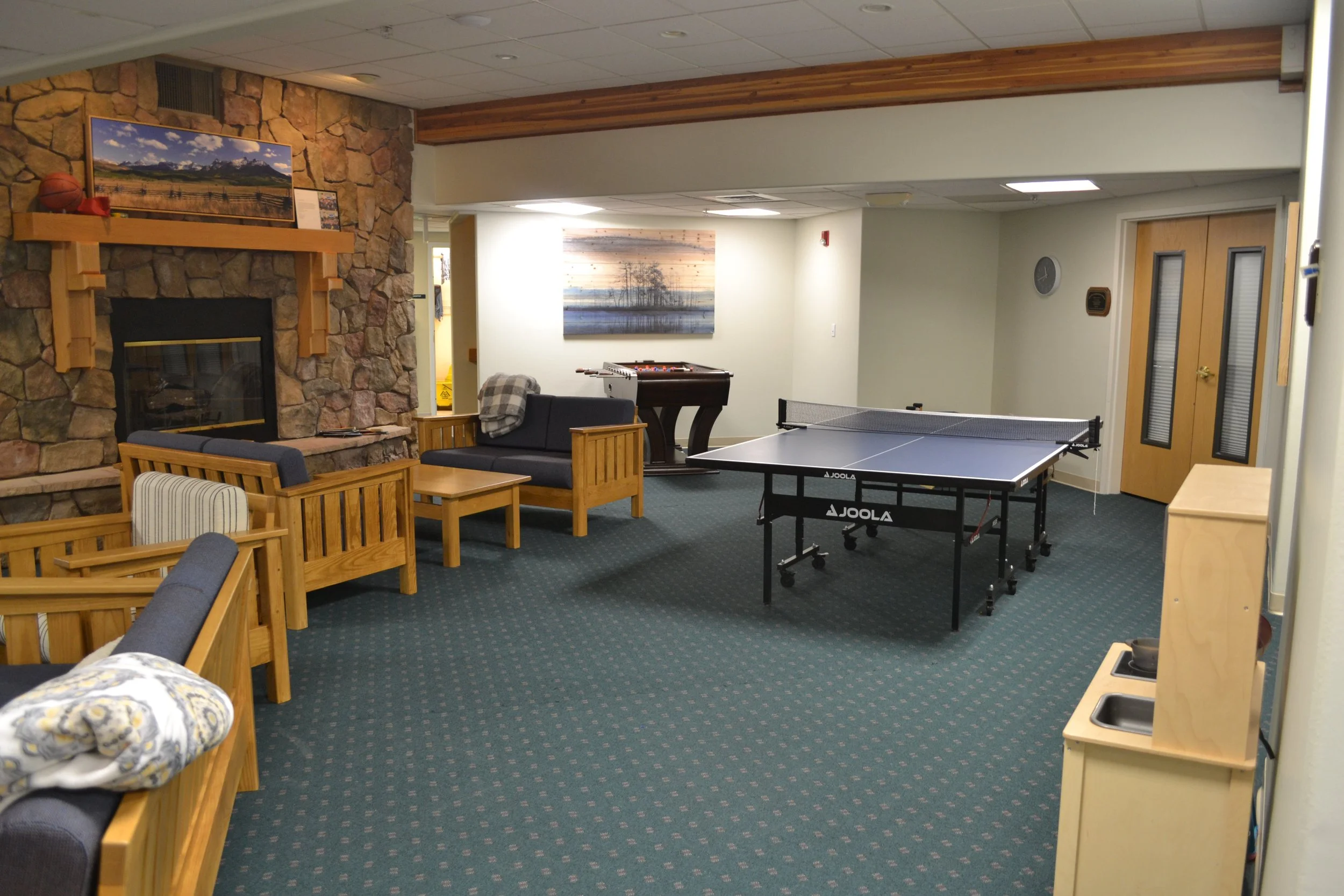 Lounge area with ping pong table, comfortable seating, fireplace, and foosball table in a recreational room with carpet flooring.