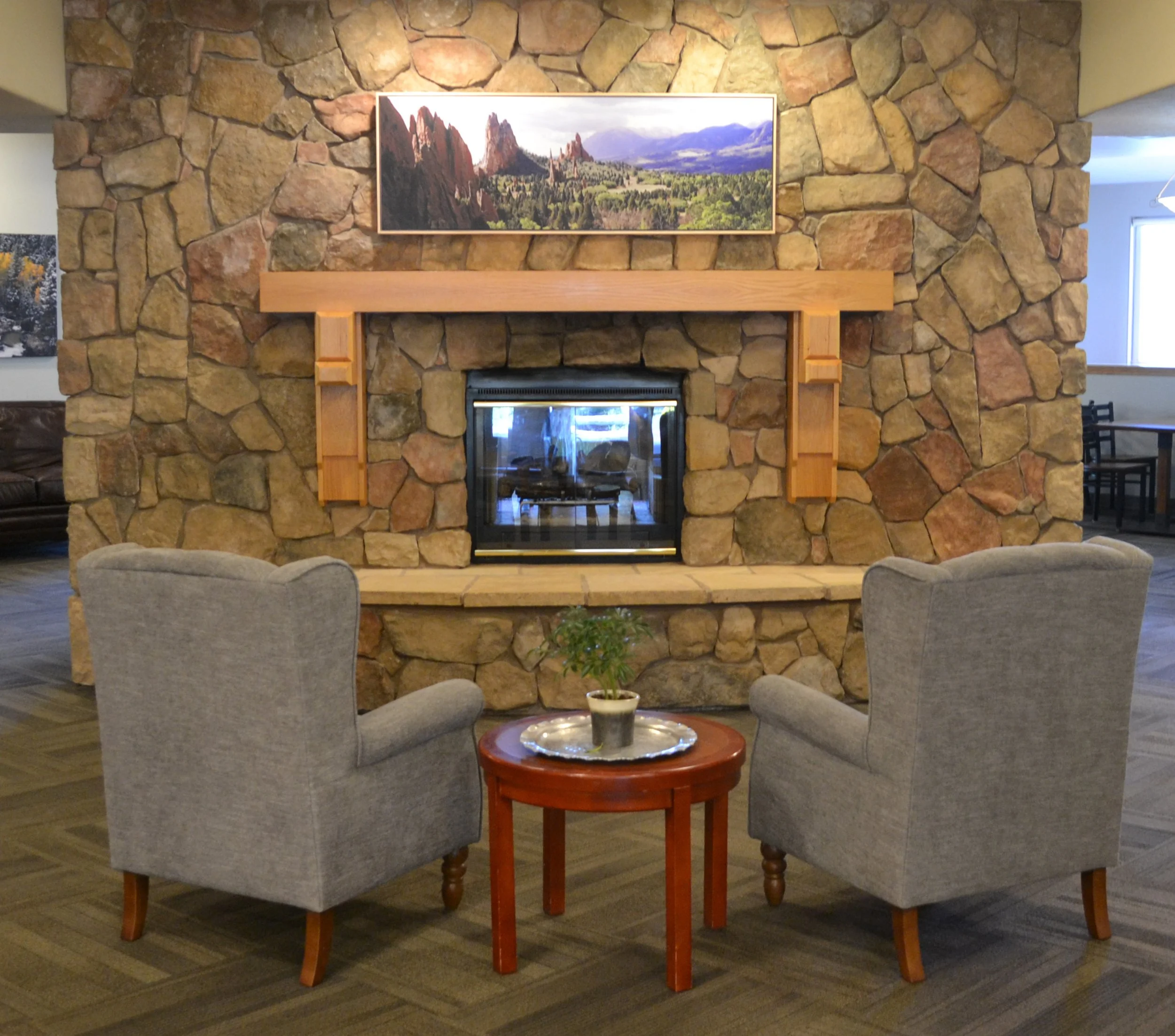 Cozy seating area with two gray armchairs facing a stone fireplace, a small round wooden table in between, and a landscape painting above the mantel.