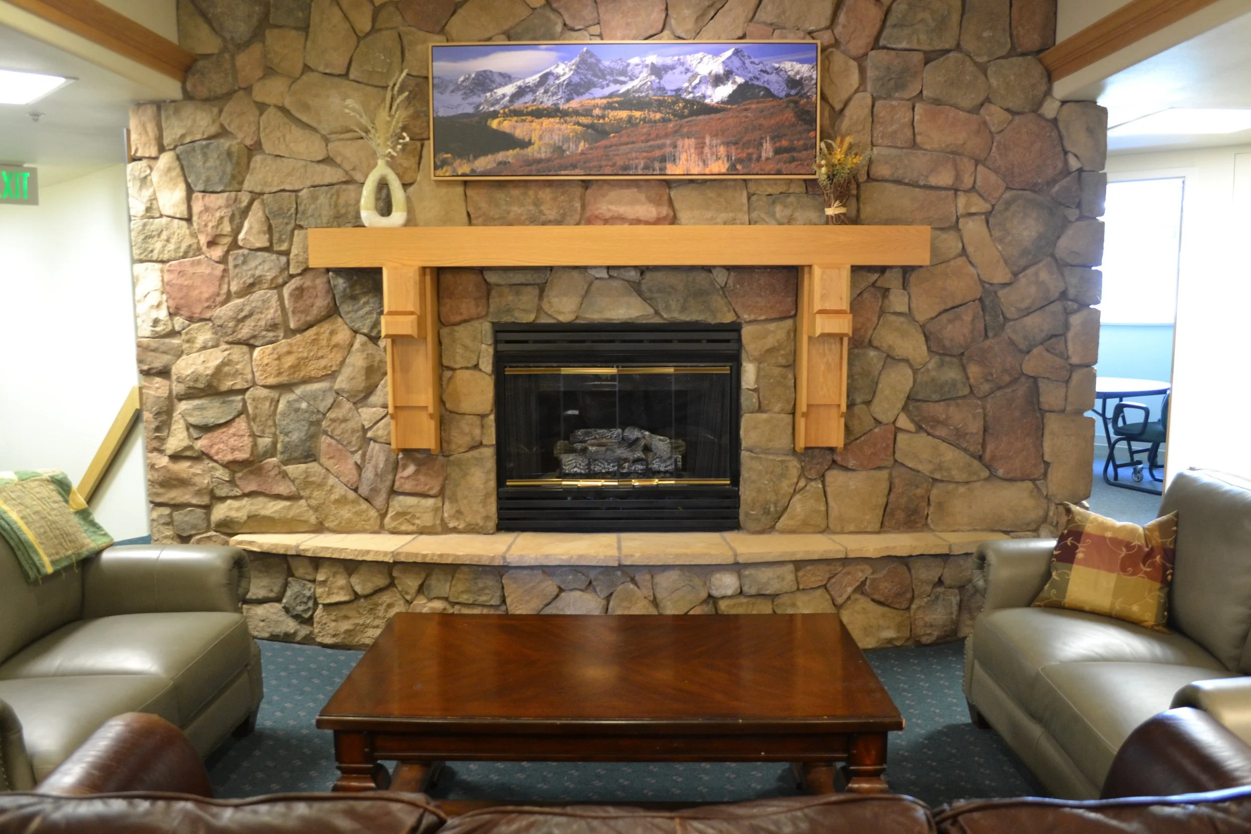 Cozy living room with a stone fireplace, wooden mantel, and landscape painting above. Two leather sofas and a wooden coffee table are placed around the fireplace.