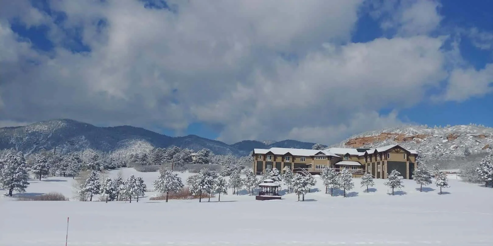 Large house in snowy landscape with trees and mountains under cloudy blue sky.