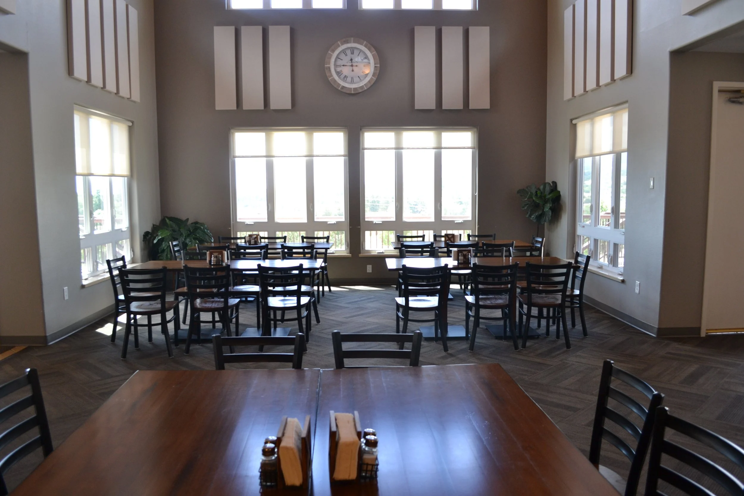 Dining room with multiple tables and chairs, large windows, a wall clock, and potted plants.
