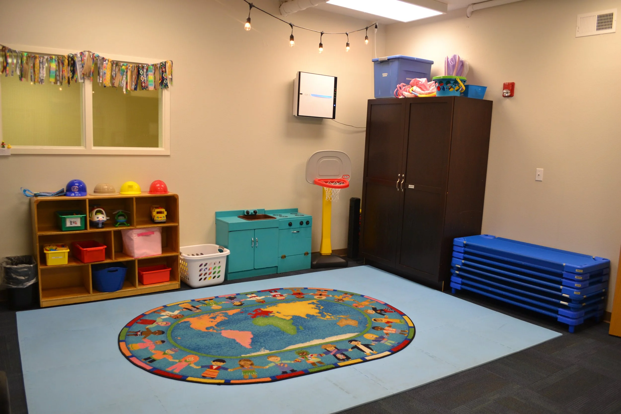 Preschool classroom with a colorful rug, toy kitchen, shelves with toys, and small basketball hoop.