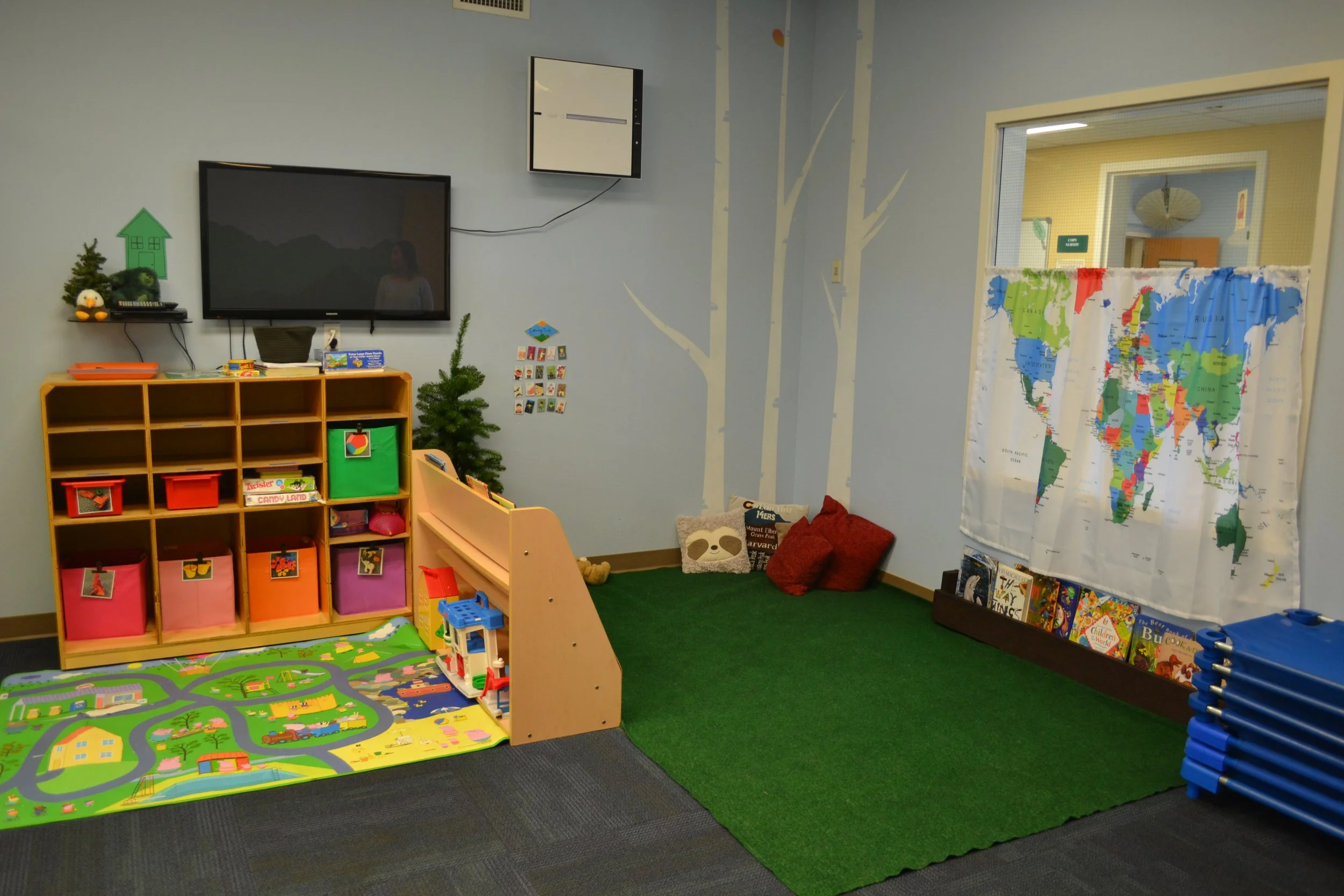 Children's playroom with storage shelves, colorful bins, a playmat, a TV on the wall, small trees, a world map curtain, pillows, books, and a stack of blue mats.