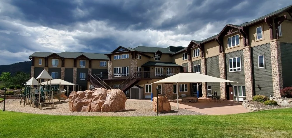 Large building with tan and green siding, adjacent playground with slides and rock climbers, patio area with shade canopy, and cloudy sky above.