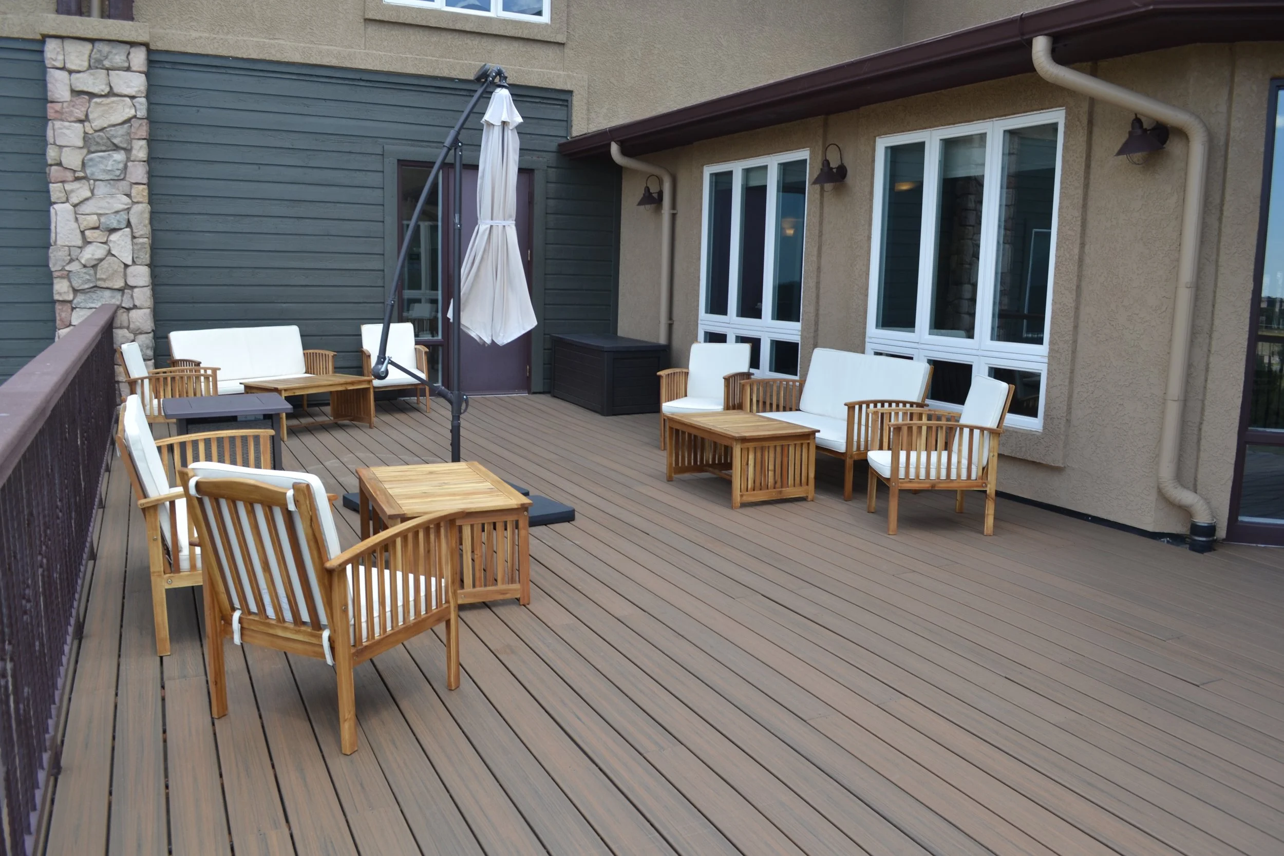 Outdoor wooden deck with seating area, wooden chairs and sofas with white cushions, a central umbrella, and tables in a residential setting.