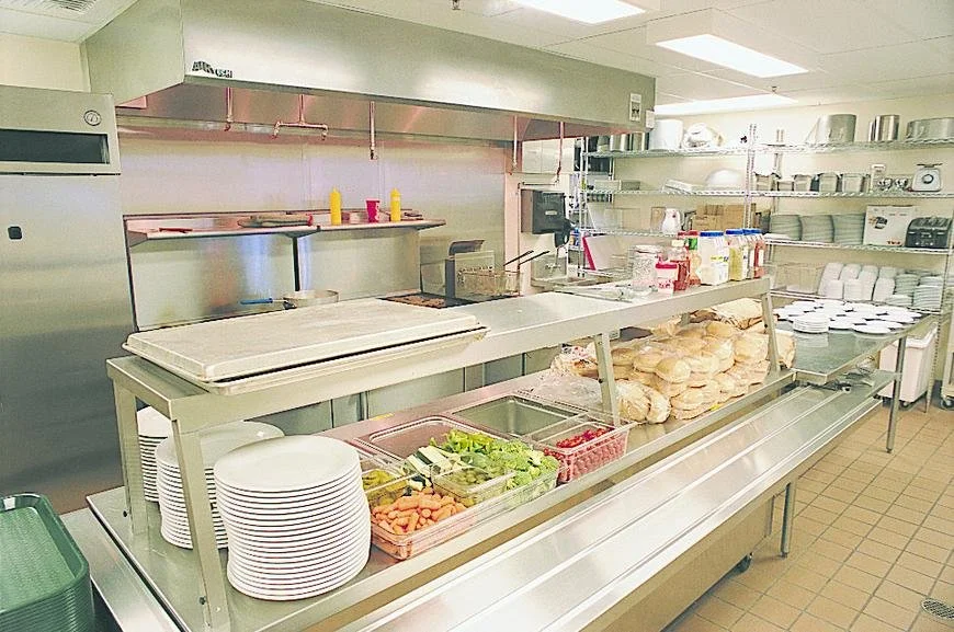 Industrial kitchen with stainless steel counters, plates, stacks of buns, vegetables in containers, condiments, and cooking equipment.