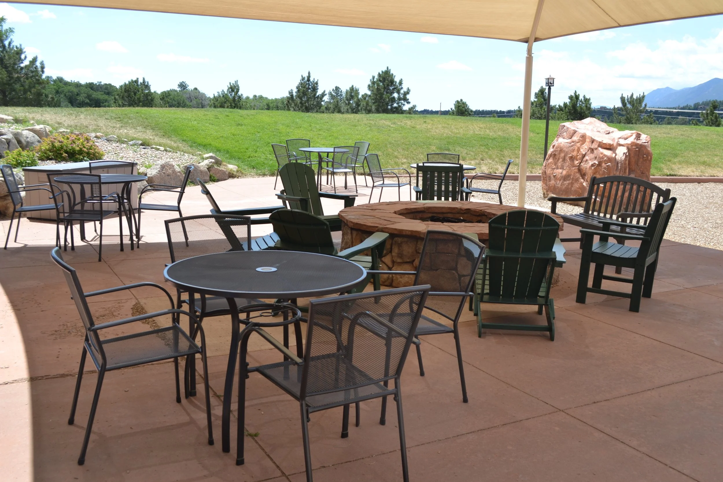 Patio with metal tables and chairs, stone fire pit, shaded area, green grass, and scenic landscape.