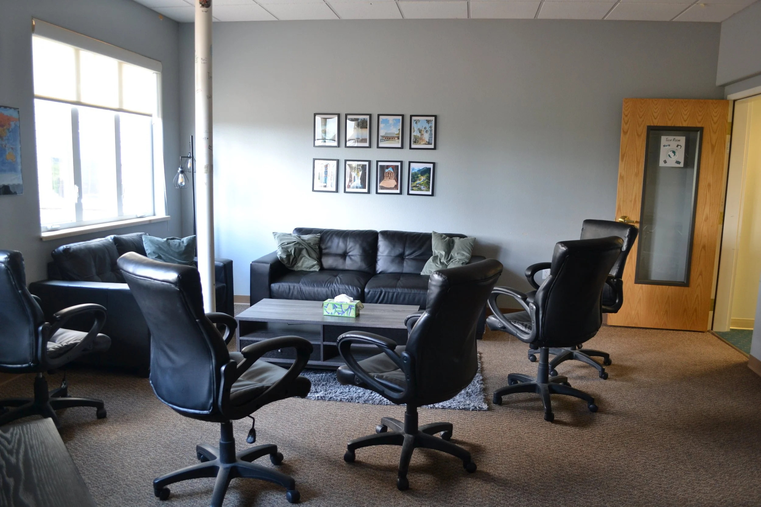 Office lounge area with black chairs and couch, a small table, framed pictures on the wall, and a window.