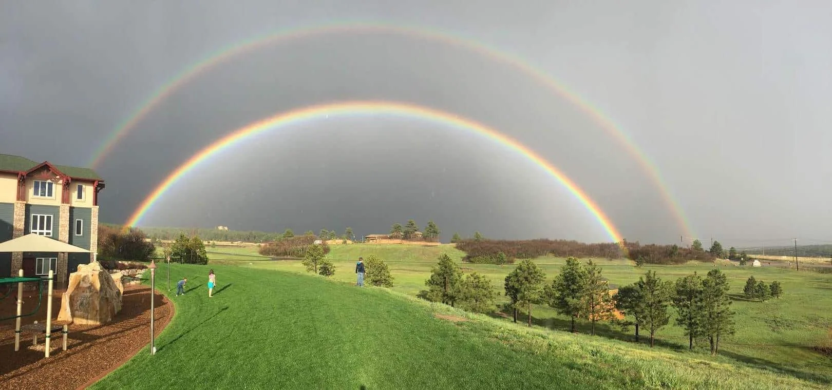 A scenic landscape showing a double rainbow over a grassy field. In the foreground, there is a playground structure and a house with children playing on the grass. The sky is overcast, enhancing the vibrancy of the rainbows.