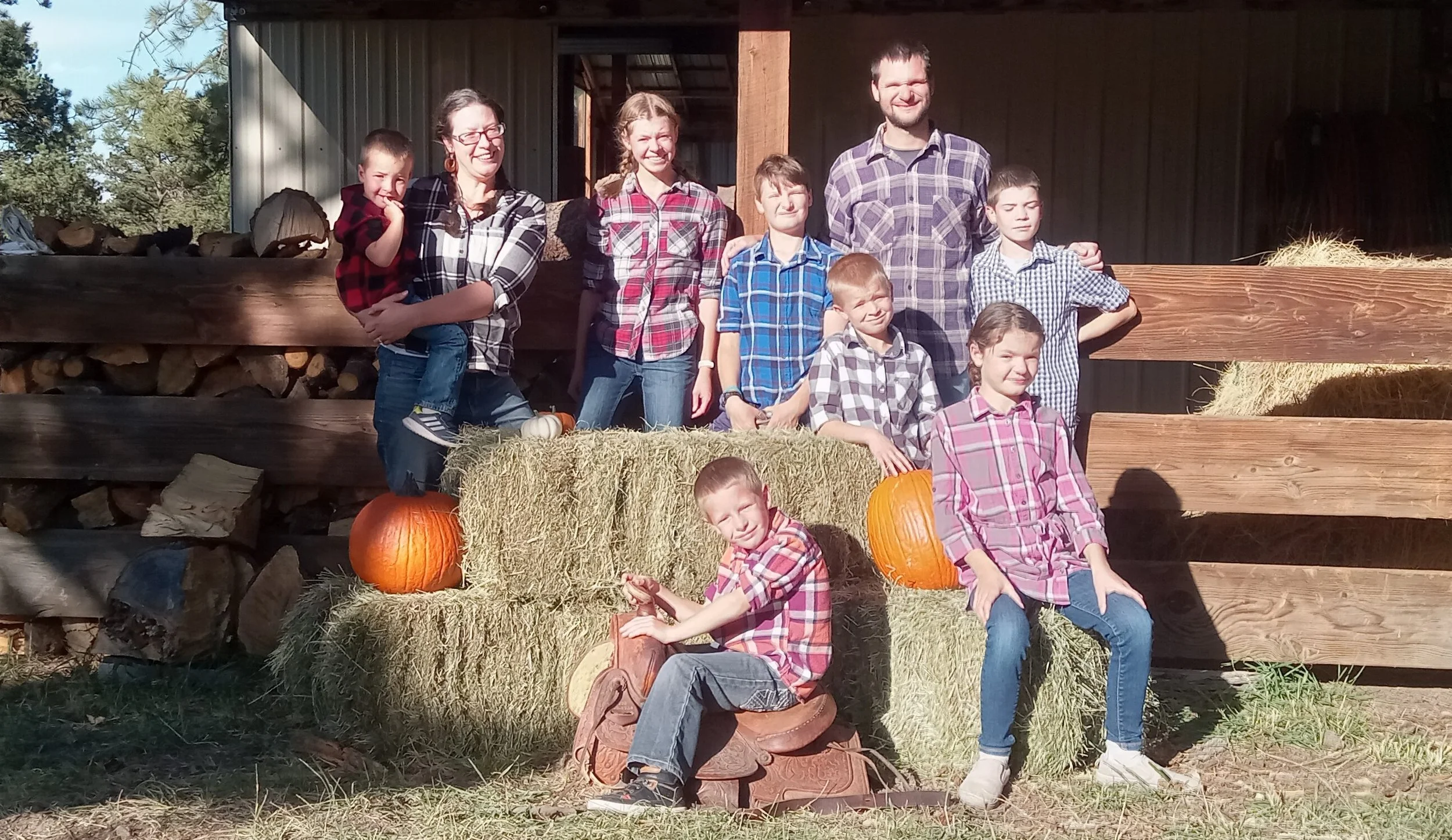 A group of people, mostly children, wearing plaid shirts, posing outdoors. They are surrounded by hay bales and pumpkins, with a wooden fence and stacked firewood in the background. One child is sitting on a saddle placed on top of a hay bale.