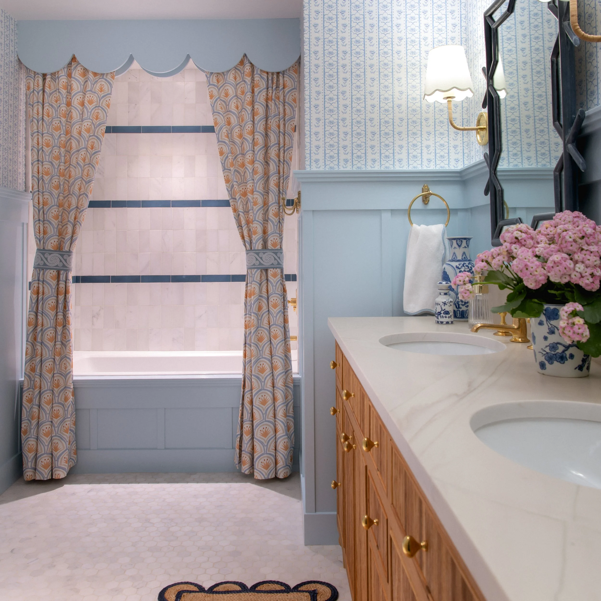A bathroom with a light blue wall, patterned curtains with orange, blue, and white designs, a white marble shower surround with a blue tile border, a white vanity with a wood finish, a sink with a white countertop, and a mirror with a black frame. There are pink flowers in a blue and white vase on the countertop, a gold wall-mounted light fixture, and a gold towel ring with a white towel.
