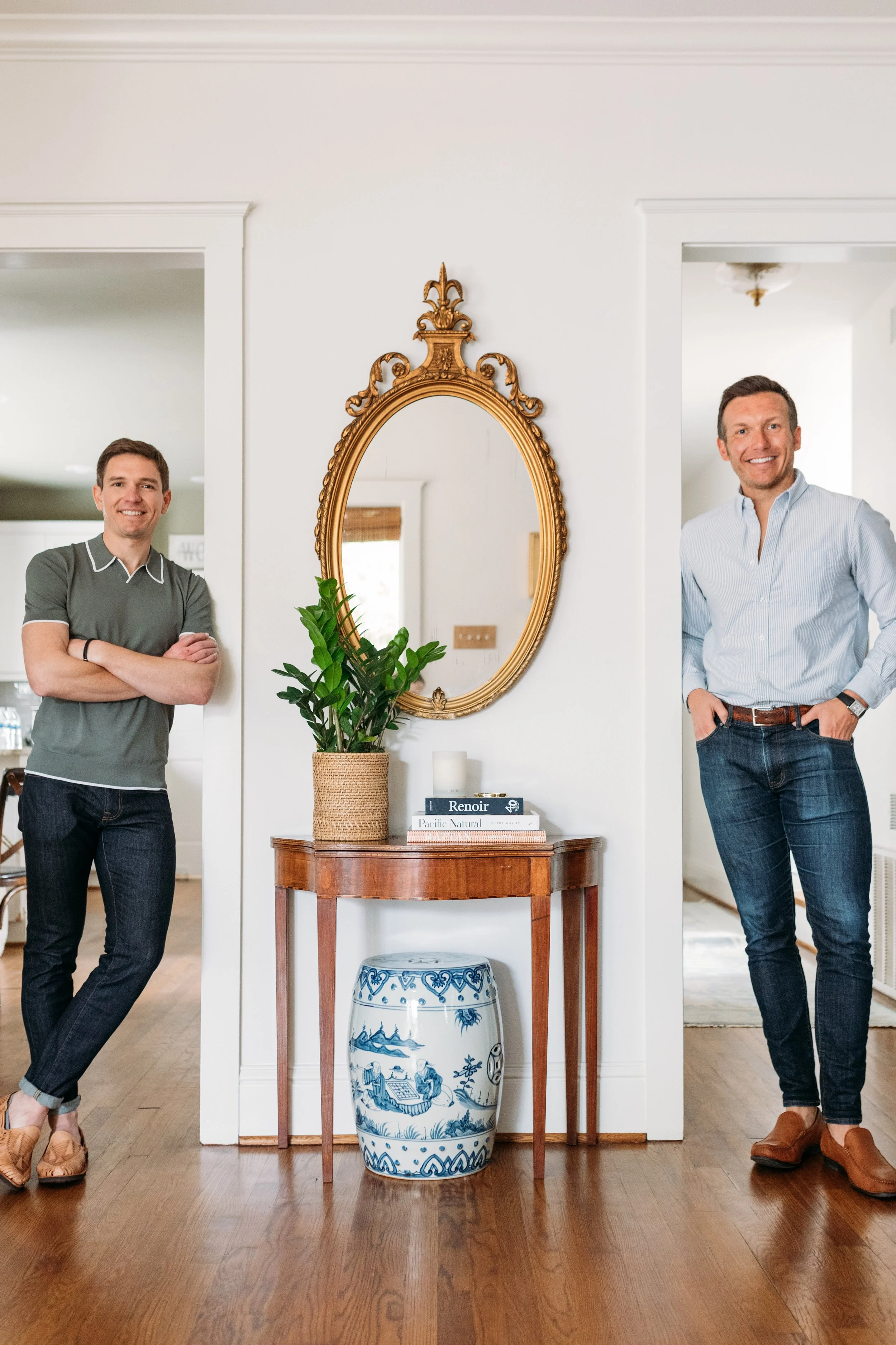 Two men stand on either side of a narrow hallway with a vintage gold mirror, a potted plant, and a blue and white porcelain urn on a wooden table in the center. The man on the left is wearing a dark green polo shirt and jeans, with arms crossed and smiling. The man on the right is wearing a light blue button-down shirt and dark jeans, with hands in pockets and smiling.