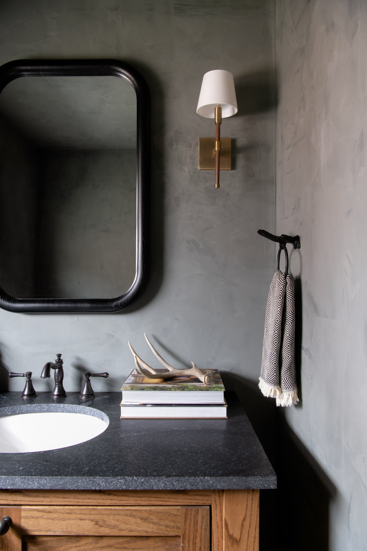A bathroom vanity with a dark countertop, a white oval sink, and three black faucets. Above the vanity is a large black-framed mirror. To the right, a beige and black patterned towel hangs from a black hook. On the countertop, there are stacked books with a deer antler on top. A brass wall sconce with a white lampshade is on the green textured wall.