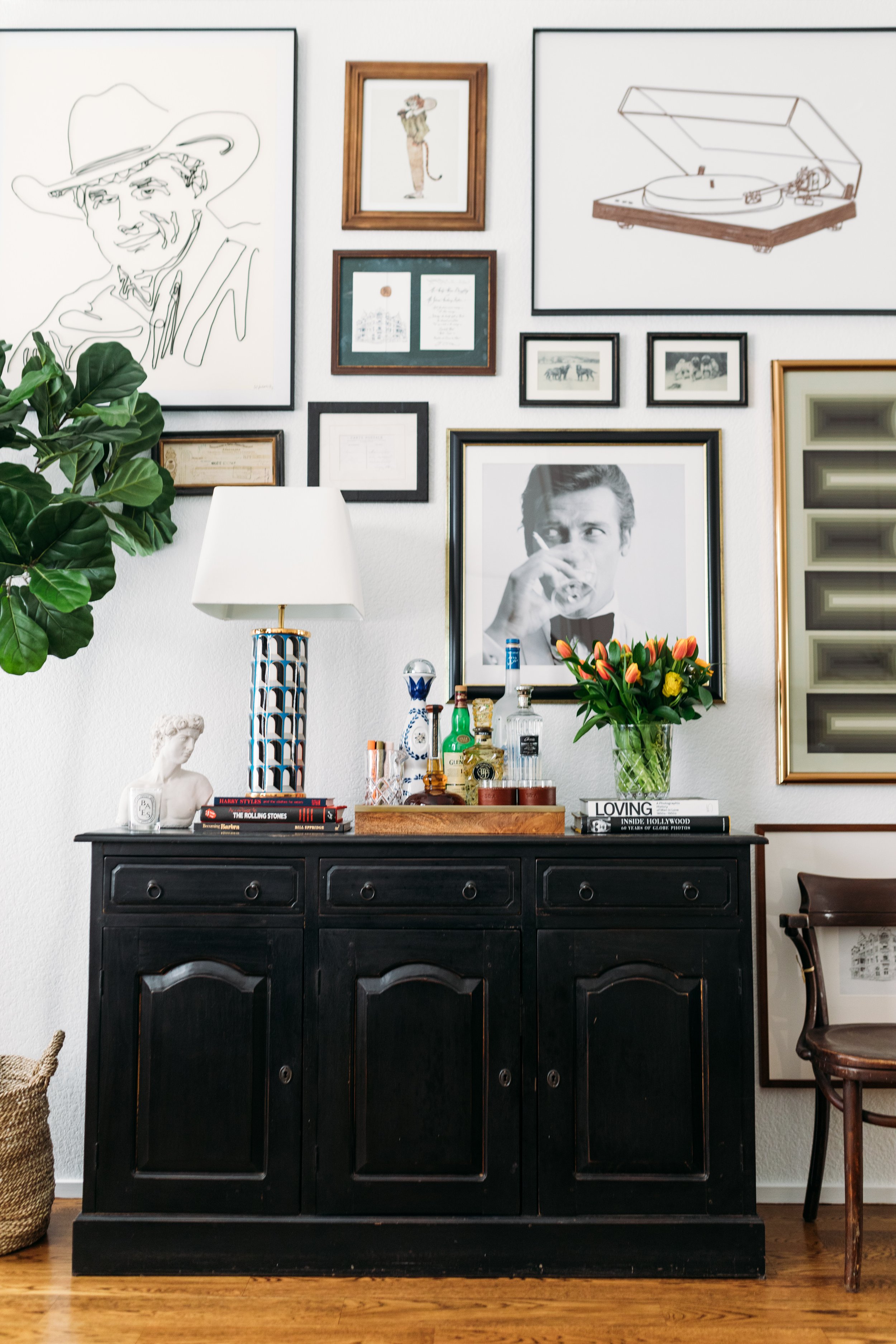 A black wooden sideboard with three drawers and cabinet doors, topped with books, a white bust sculpture, a blue and white vase, bottles, and a glass container. On top of the sideboard is a flower vase with a bouquet of yellow and orange flowers, a white lamp, and various small items. The wall above features multiple framed artworks, including a large black and white photograph of a man drinking, a line drawing of a smiling person in a hat, a drawing of a person with a hat and a cat tail, a vintage postcard, and several smaller pictures. There is greenery on the left side of the image and a wooden chair on the right.