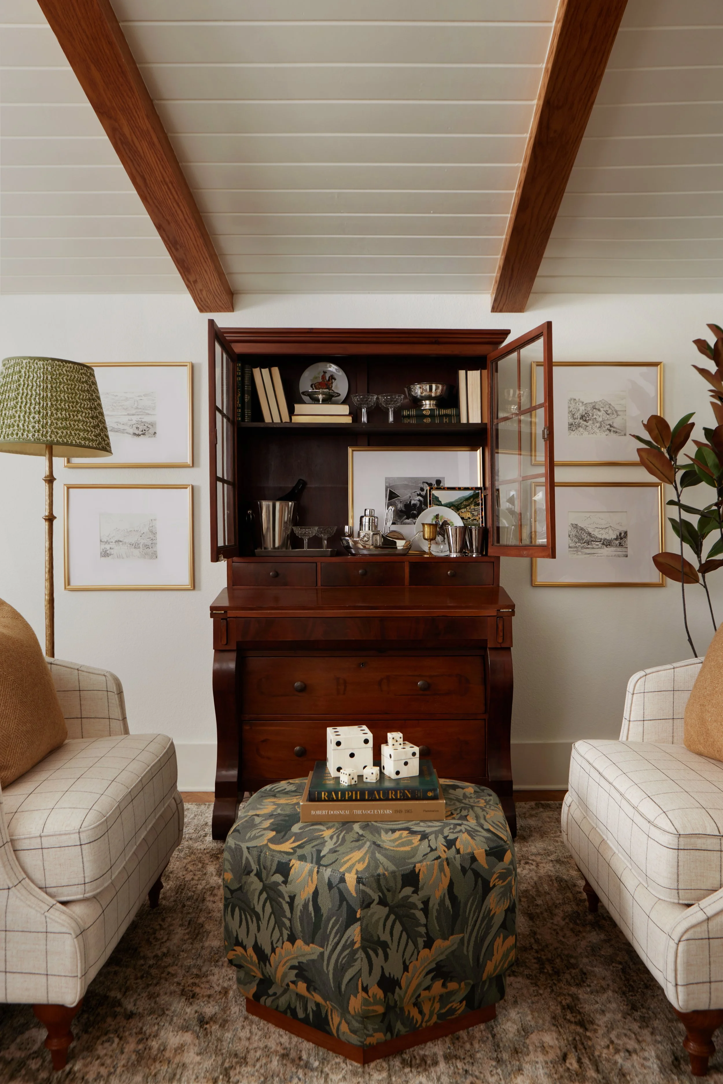 Sitting room with two plaid armchairs facing each other, a patterned ottoman with dice and books in the center, a wood china cabinet with glass doors displaying glassware and books, and framed artwork on the wall.