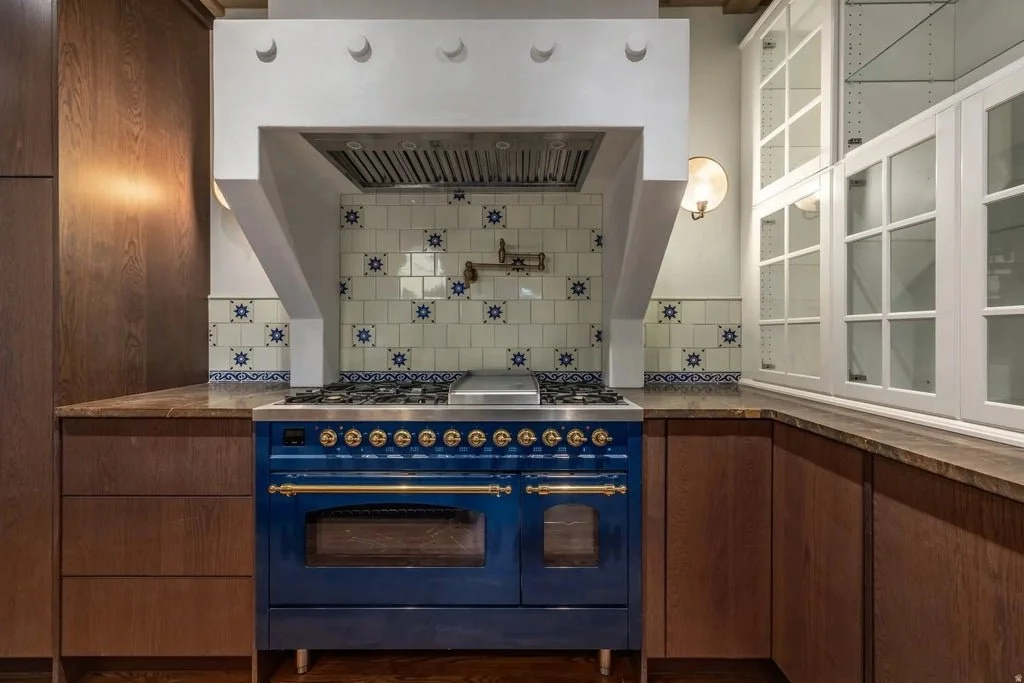 A kitchen with a blue oven, white tiled backsplash with blue accents, wooden cabinets, and a glass-fronted cabinet on the right.