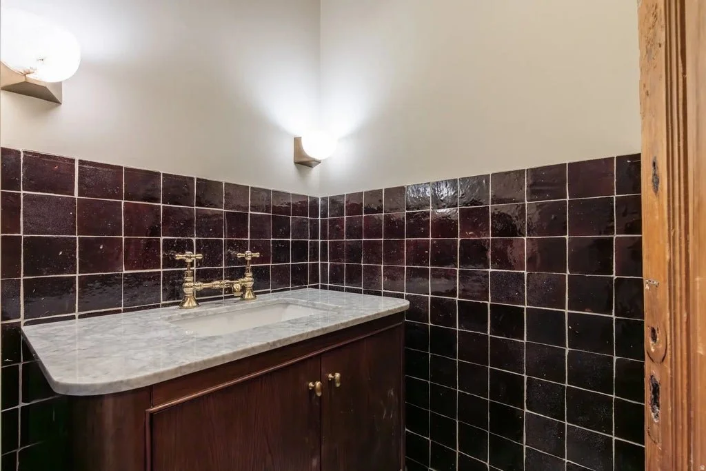 A bathroom corner with a wooden cabinet under a marble countertop sink, brass faucet, black tiled walls, and wall-mounted light fixtures.