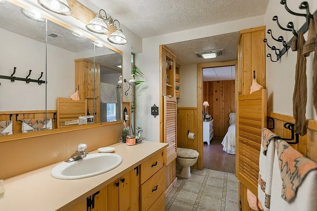 Bathroom with a wooden vanity, mirror, and sink; toilet; towels hung on hooks; view into the bedroom with a bed and nightstand.