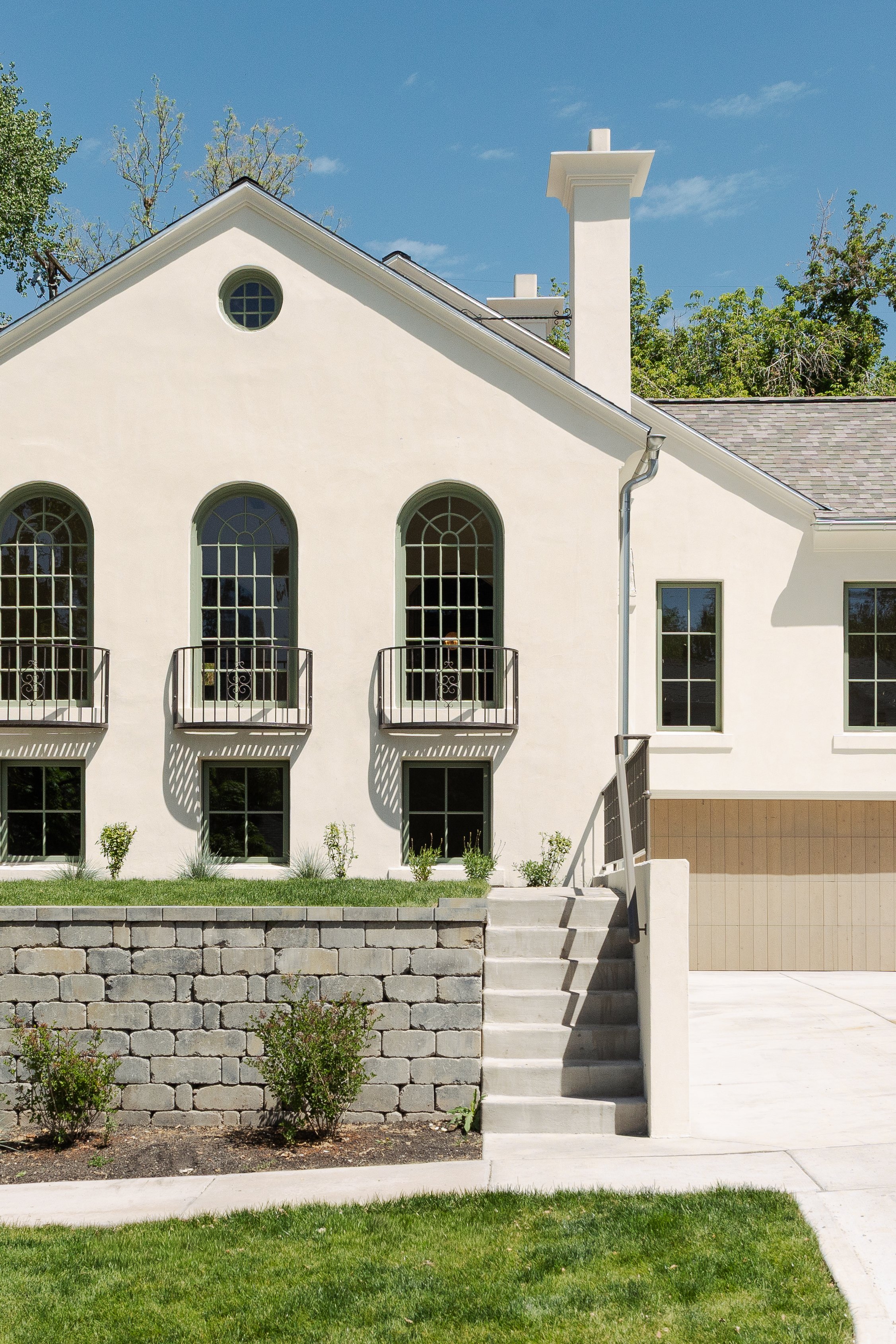 White house with arched windows, small balconies, green lawn, and stone retaining wall.