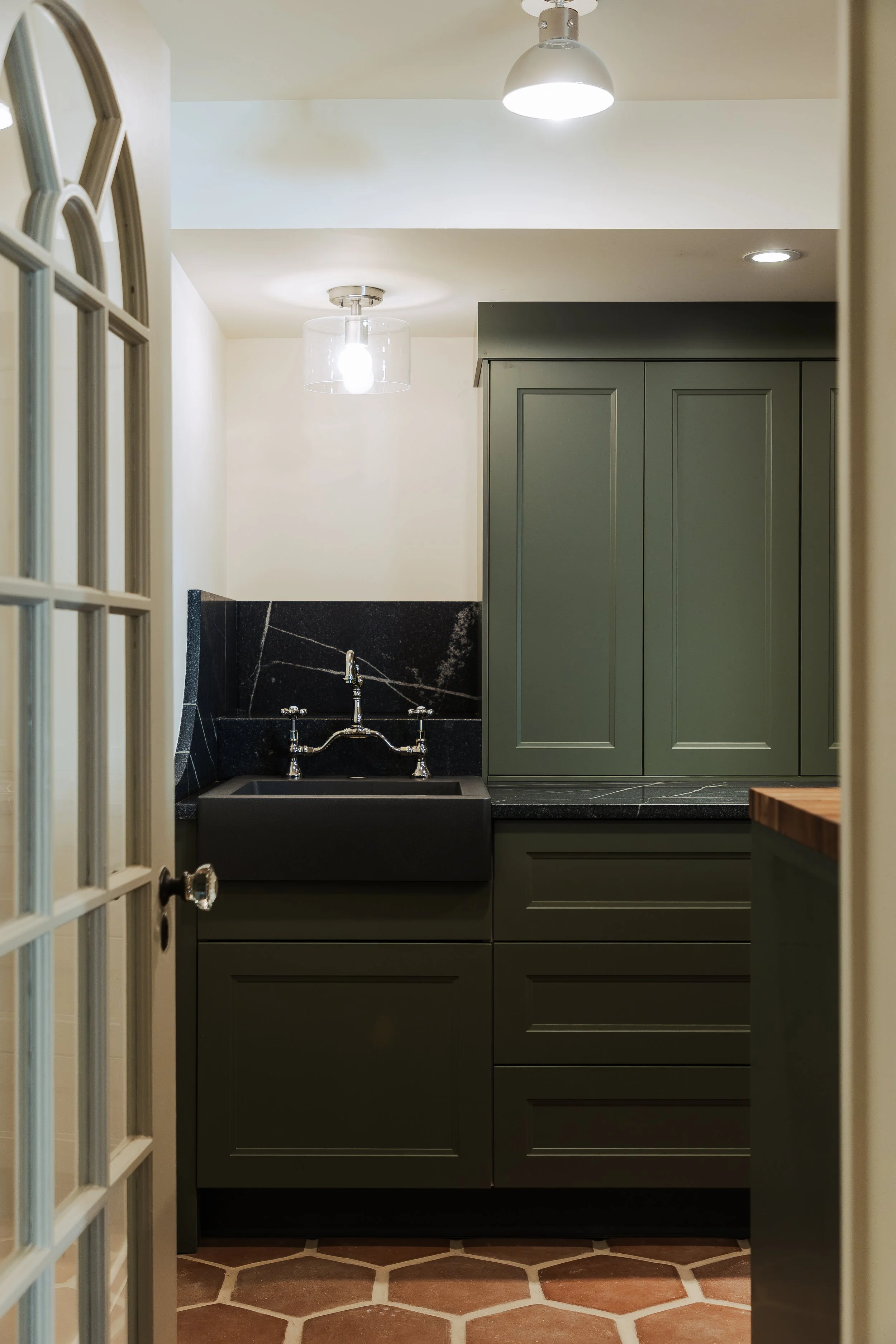 A small laundry room with a black sink, dark green cabinets, and hexagonal terracotta tile flooring. The ceiling has modern pendant and recessed lighting fixtures.