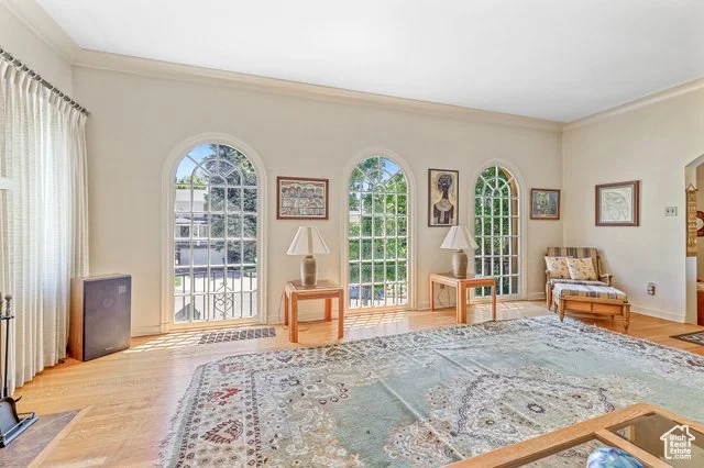 A spacious living room with hardwood floors, large arched windows, and a decorative area rug. There are two wooden side tables with lamps, a framed painting between the windows, and a small seating area with a patterned armchair. The room is well-lit