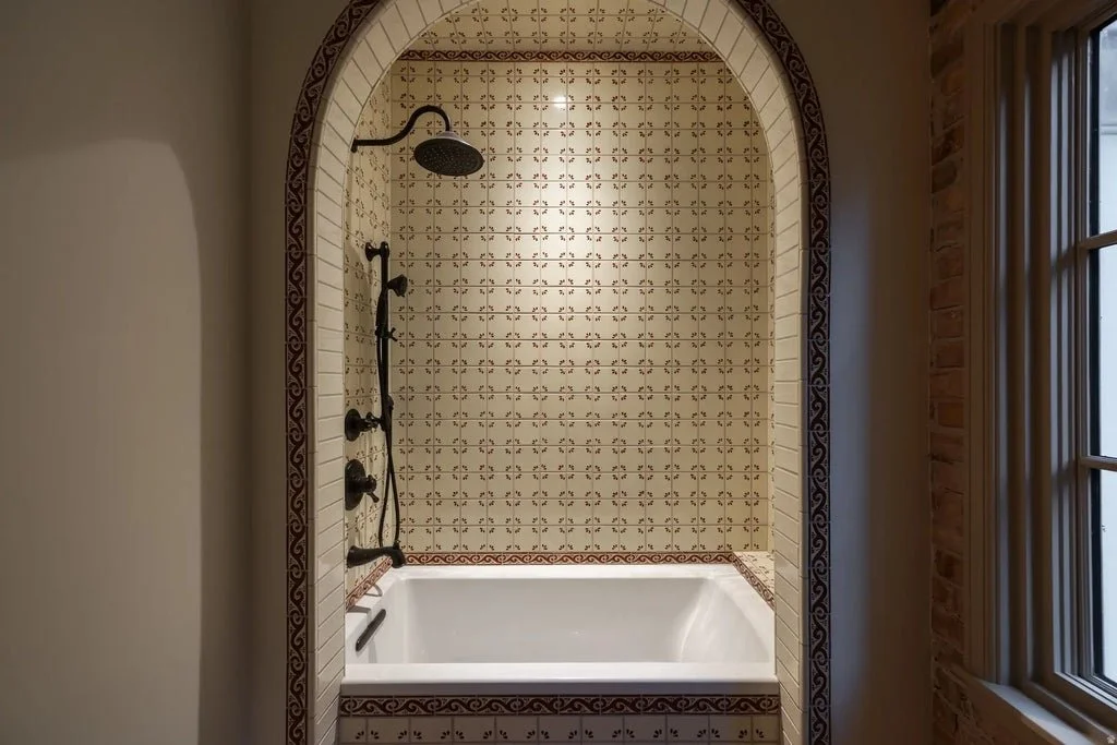 Bathroom with a built-in bathtub, shower head and fixtures, surrounded by decorative tiles and an arched entryway, with a window to the right.