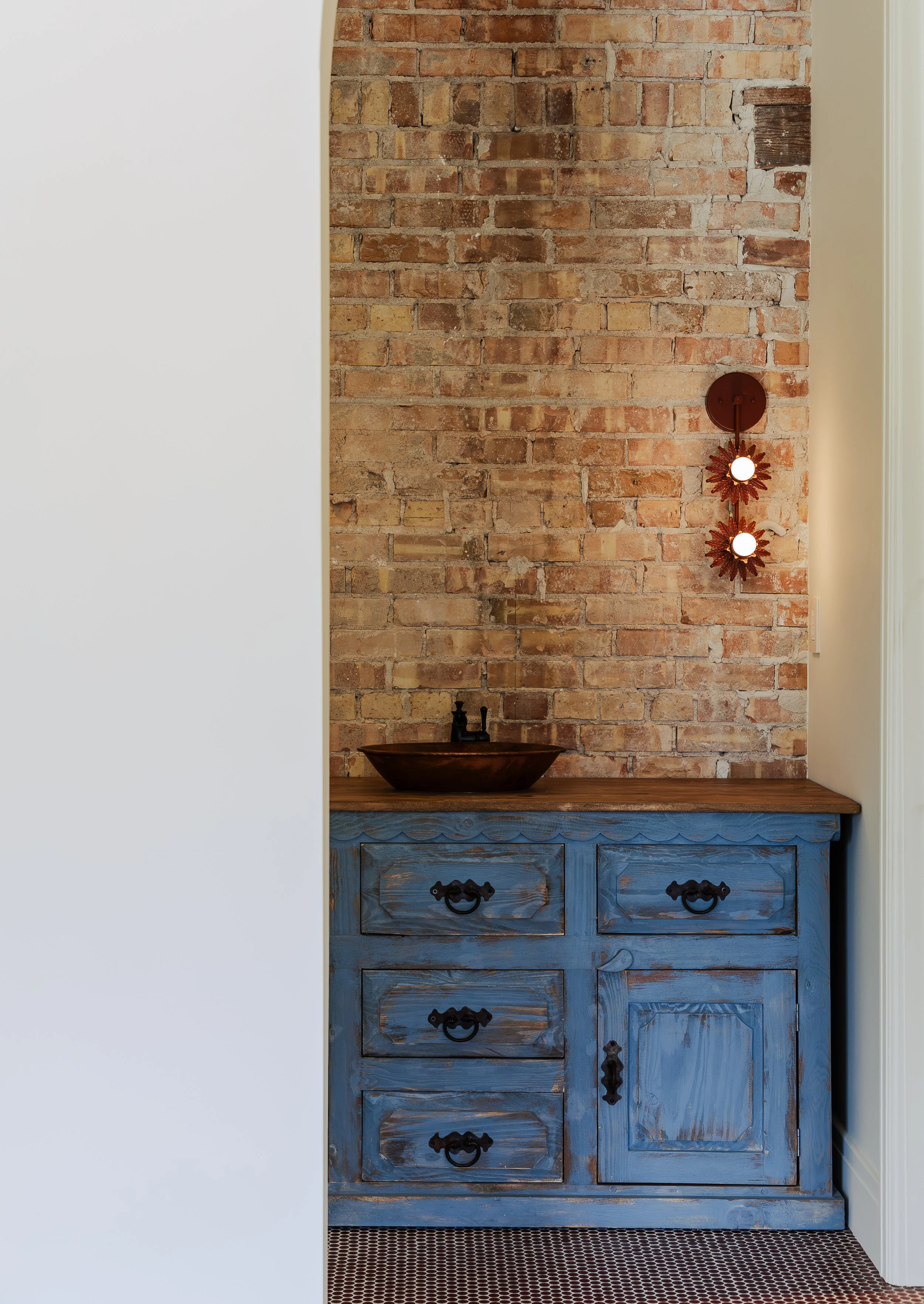 View of a rustic blue wooden cabinet with black hardware, placed against an exposed brick wall with a wooden basin and a modern light fixture above it.