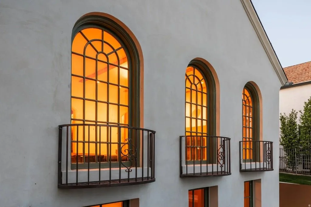 The exterior of a building with three arched windows, each with a small black metal balcony, illuminated by warm orange interior lighting.