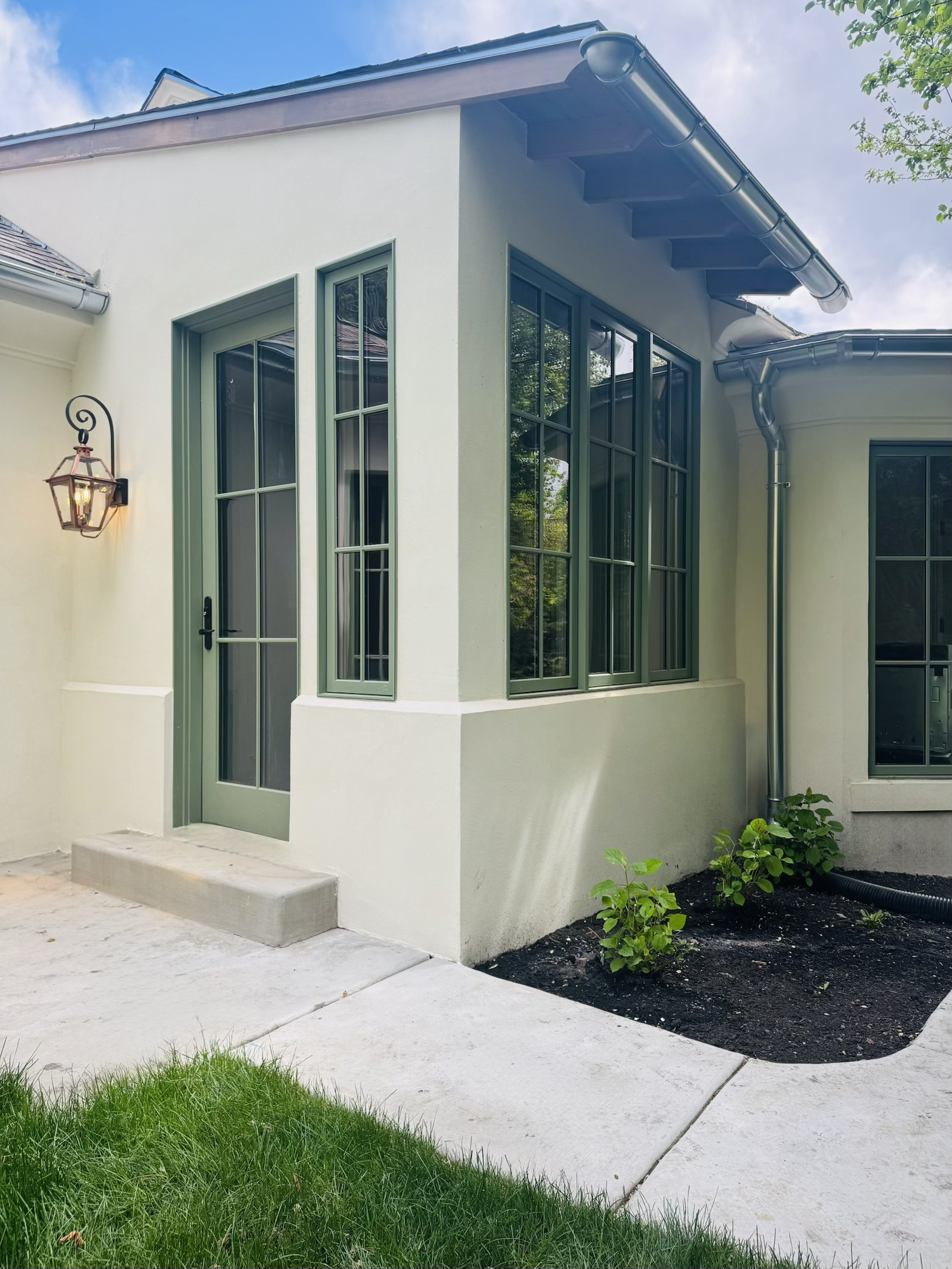 Exterior view of a house with light-colored walls, large green-framed windows, a small front step, a black door handle, a wall-mounted lantern, and a garden with small green bushes, a concrete sidewalk, and a grassy area.