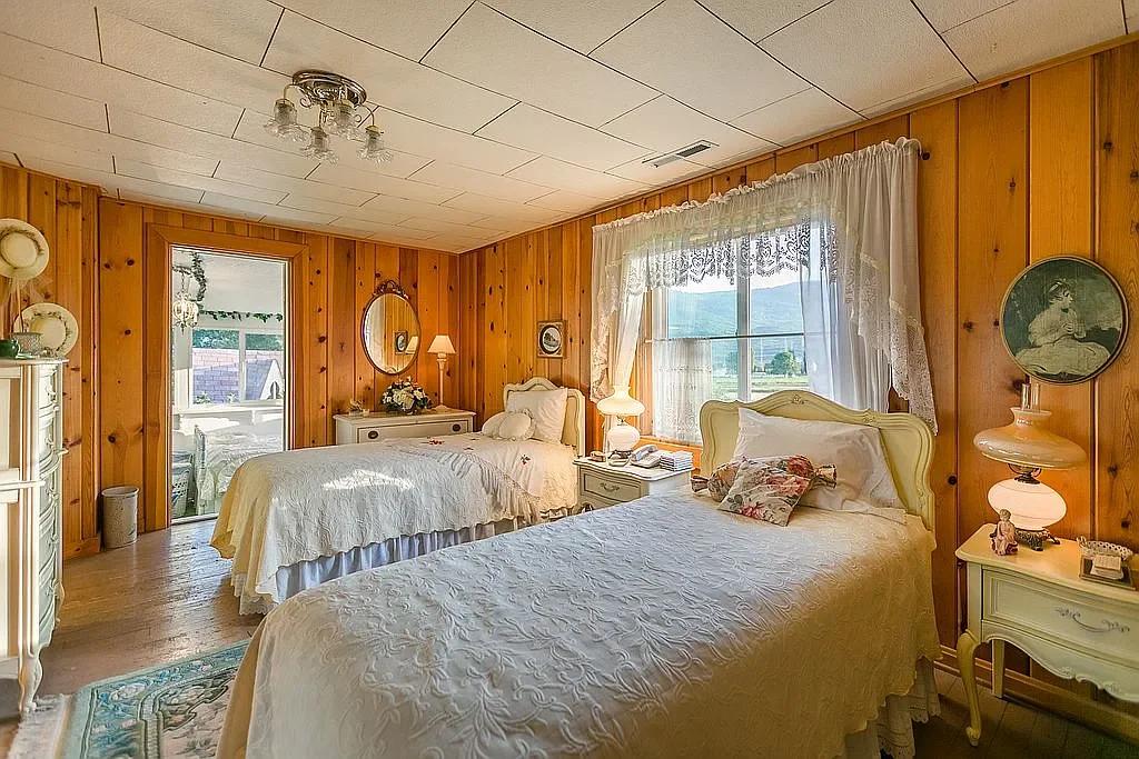 A bedroom with wood-paneled walls, two single beds with white bedding, and vintage furniture. There's a window with lace curtains, a mirror on the wall, and a door leading outside. The room has a cozy, rustic feel.