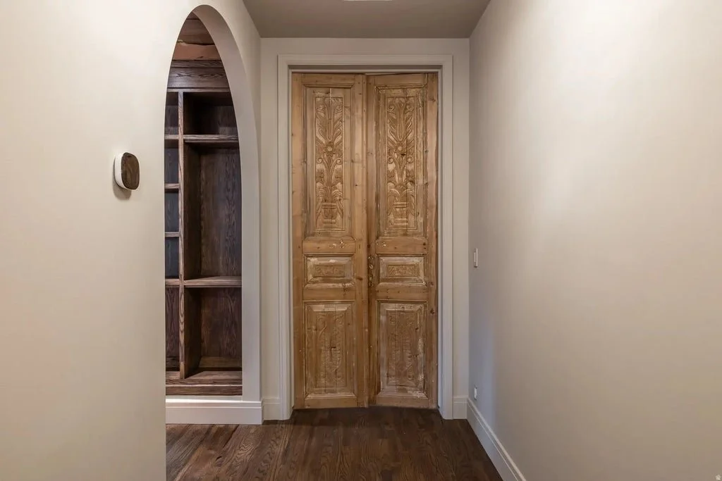 Wooden double doors in a hallway with beige walls and hardwood flooring, with a built-in wooden shelving unit on the left side.