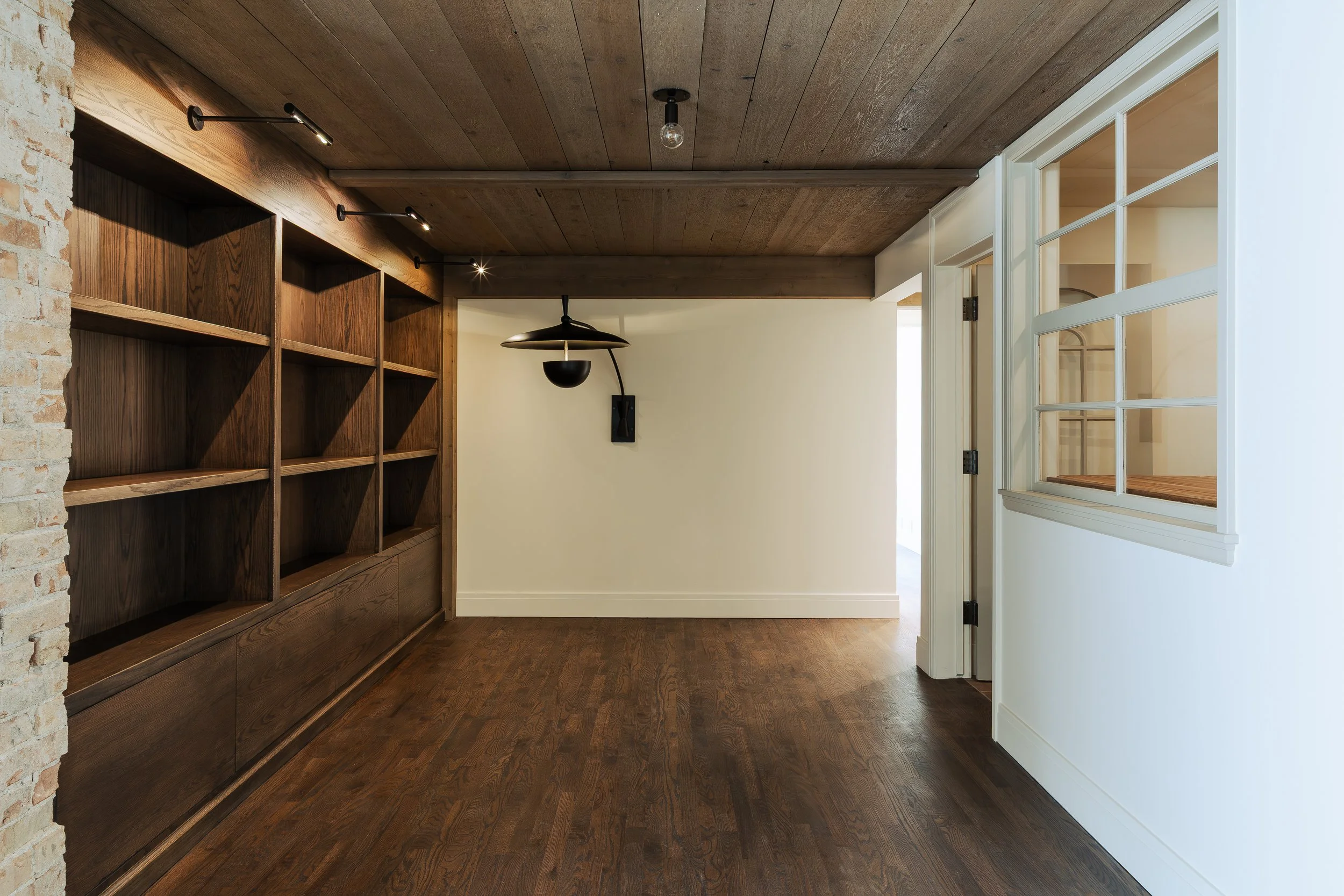 Interior of a room with wooden flooring and a wooden wall with open shelves on the left, a white wall with a window on the right, a hanging light fixture, and track lighting on the ceiling.