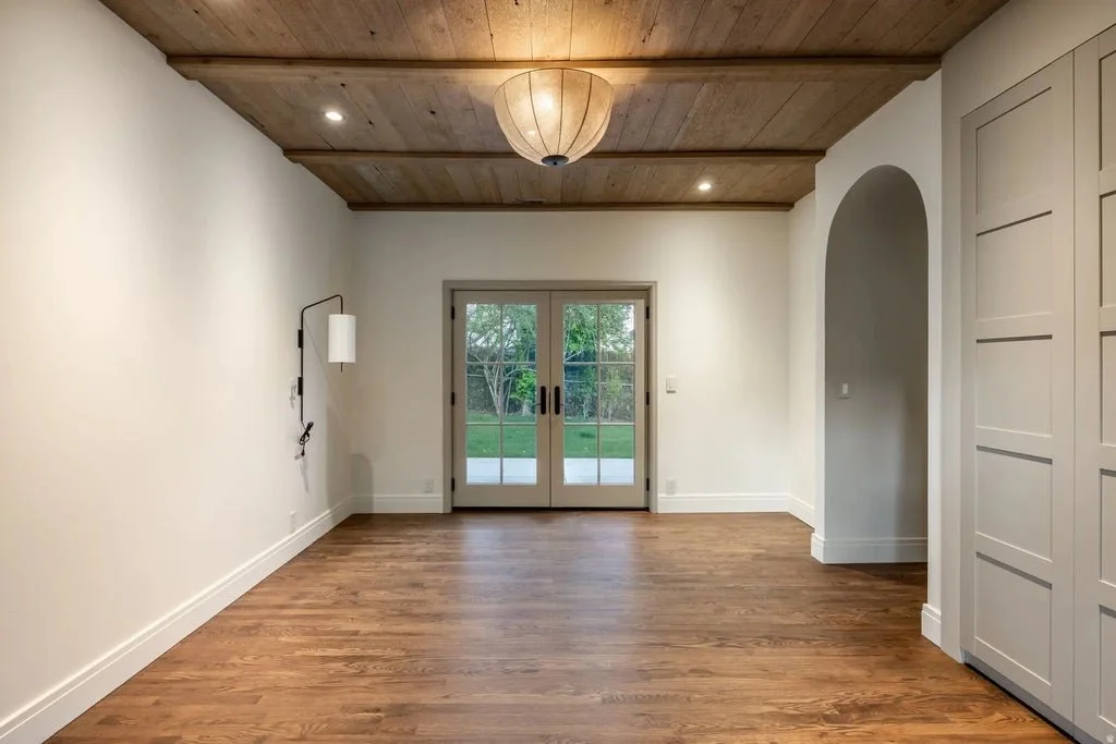 Empty room with wooden flooring, white walls, and a wooden ceiling. Features a ceiling light, a wall-mounted lamp, and glass double doors leading outside to a grassy yard.