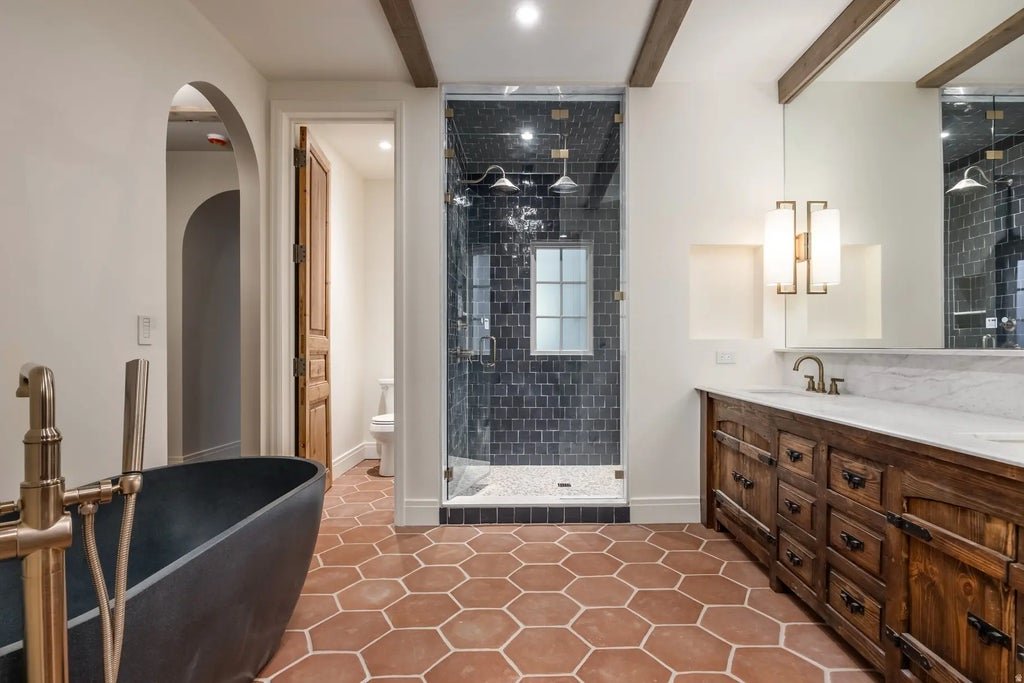 Modern bathroom with terracotta hexagon floor tiles, a black bathtub, a wooden vanity with a marble countertop, a large mirror, wall-mounted light fixtures, and a glass-enclosed shower with black subway tiles.