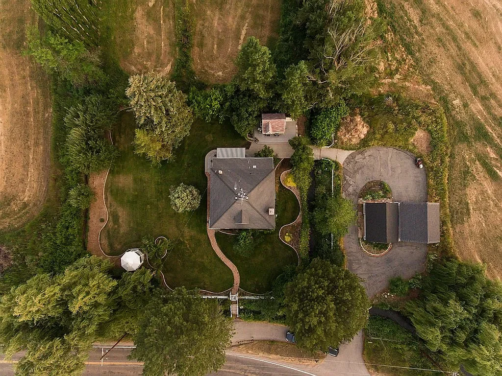 Aerial view of a house with a curved walkway, surrounded by trees, a shed, and a driveway on a spacious property with open fields.