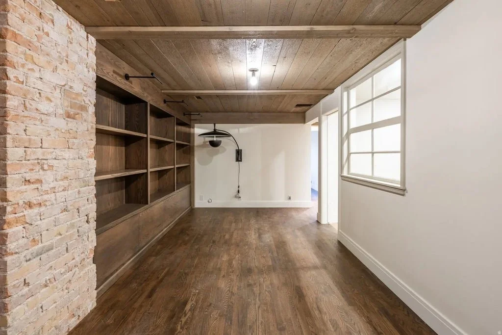 Empty room with wooden floors, a brick wall on the left, built-in wooden shelves on the left wall, and a window on the right wall.