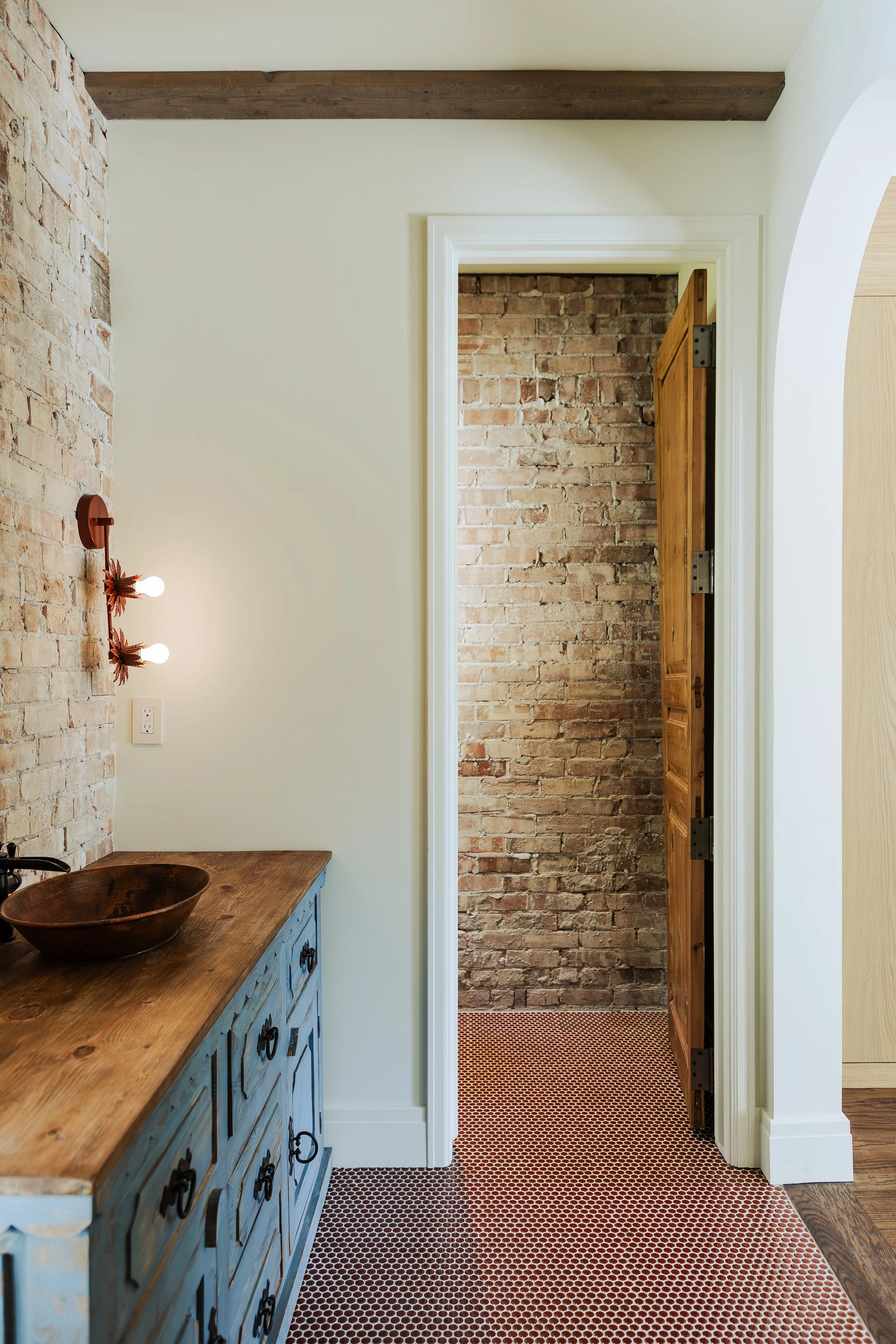 Interior view of a room with a rustic decor, featuring a blue wooden sideboard with drawer and door cabinet, a wooden bowl on top, exposed brick wall, and a doorway with wooden door leading to a space with brick wall and patterned red flooring.