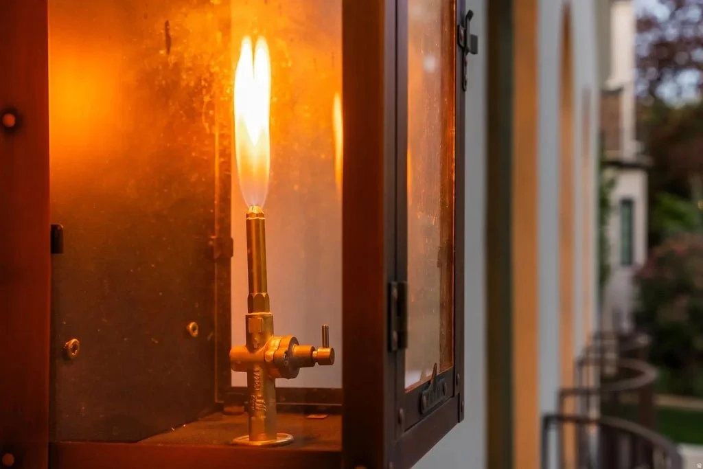 A close-up of a Bunsen burner with a lit flame inside a heated metal box attached to an electrical outlet on an exterior wall.