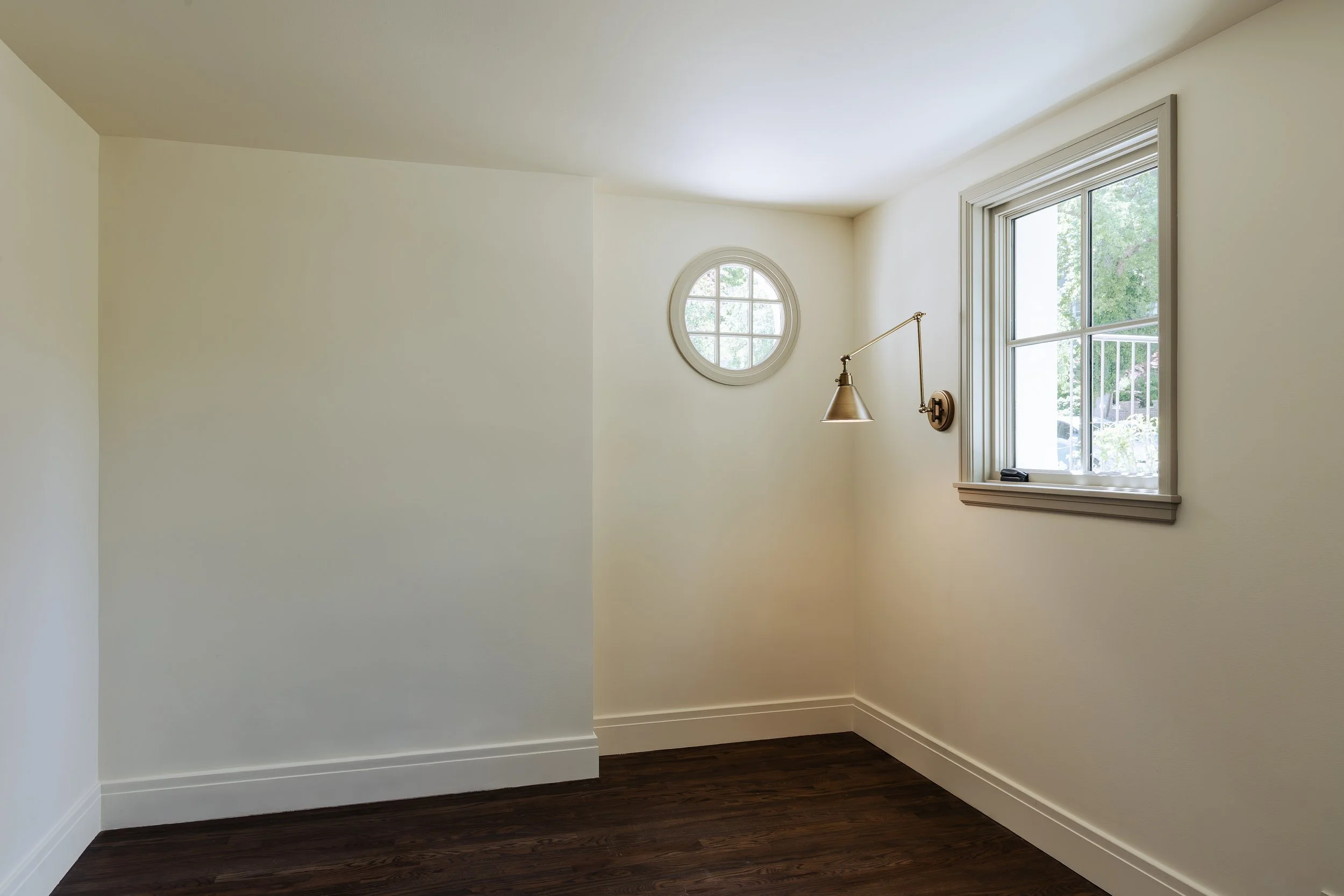 Empty room with white walls, dark hardwood floors, a large window with a window seat, and a round window on the adjacent wall.