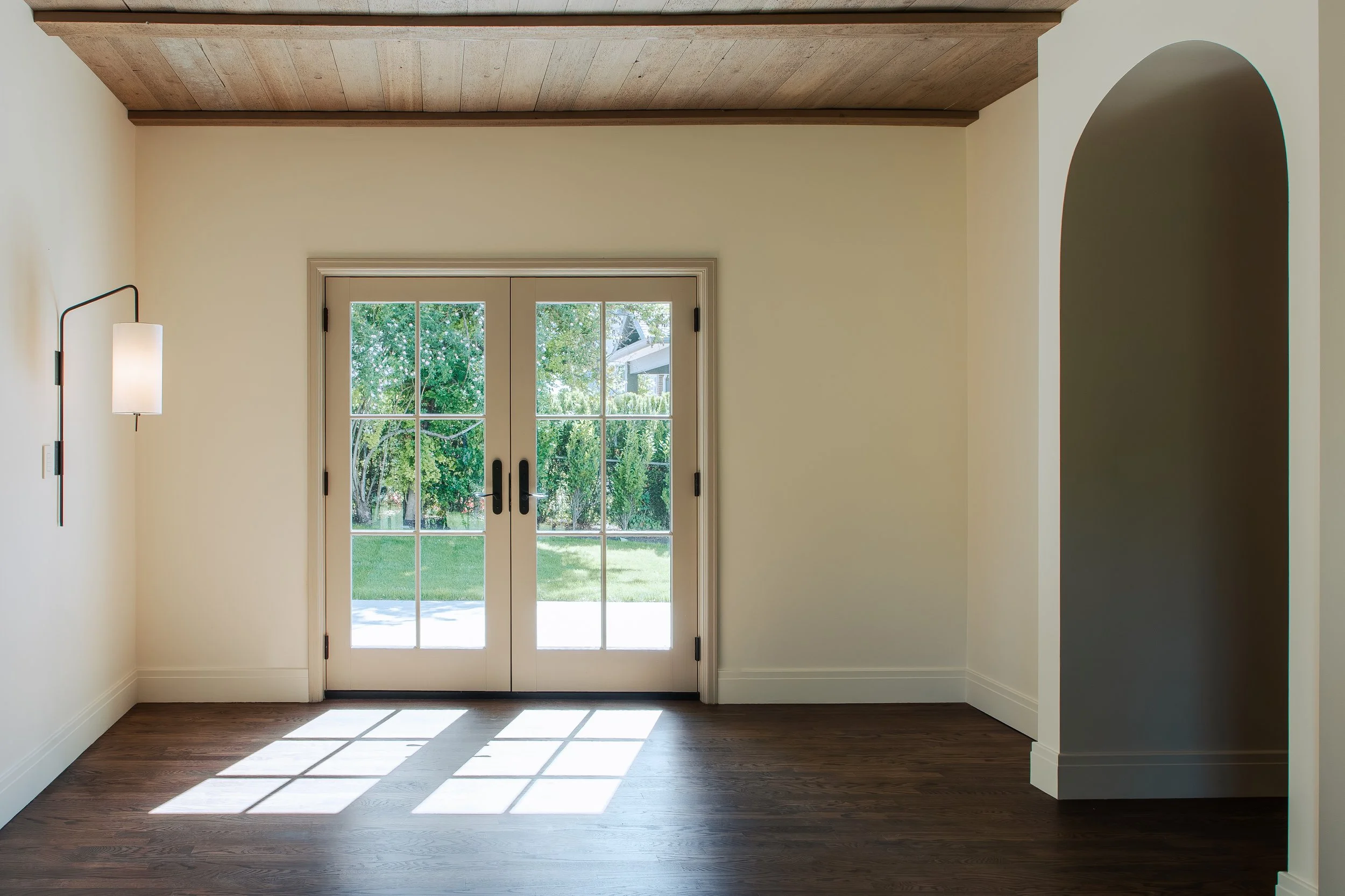 A room with a wooden ceiling, cream-colored walls, and dark wooden flooring. Double glass doors lead outside to a yard with green grass and trees. A wall-mounted lamp with a white shade is on the left wall, and an arched opening is on the right side.