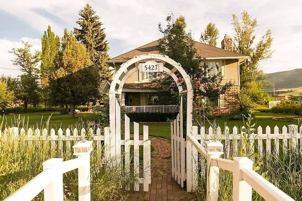 A white picket fence gate with a decorative archway leading to a large two-story house surrounded by greenery and trees.